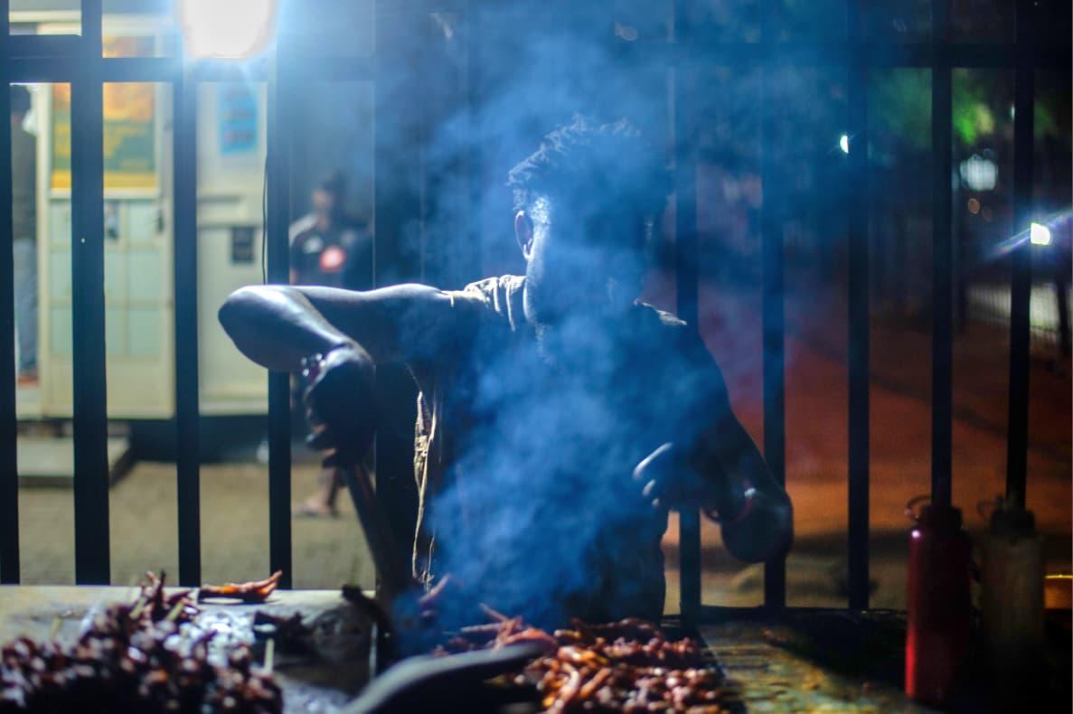 Sizzling Streets: The Night Vendor Swirling smoke and bright overhead lighting surround a street cook preparing food at a bustling night stall.