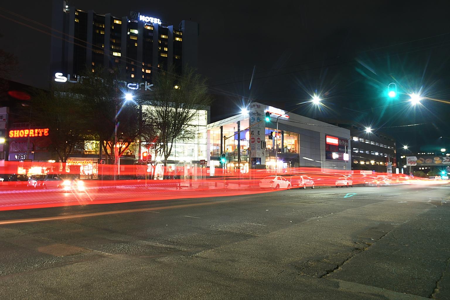Intersections at Twilight: Crossing the Street: Red traffic signals and glaring car headlights slice through the dusk as a pedestrian pulls a crate across a quiet intersection. 