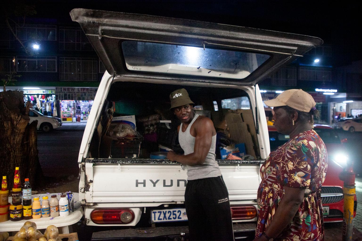 The Street-side Merchant:  A local vendor stands with his back to the open trunk of his vehicle, engaging in conversation with a customer. 