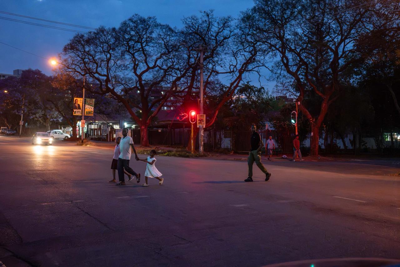 Dusk Crossing: Street at Dusk:  Pedestrians, including a family holding hands, cross an illuminated street framed by jacaranda trees and red traffic signals under a deep twilight sky.   