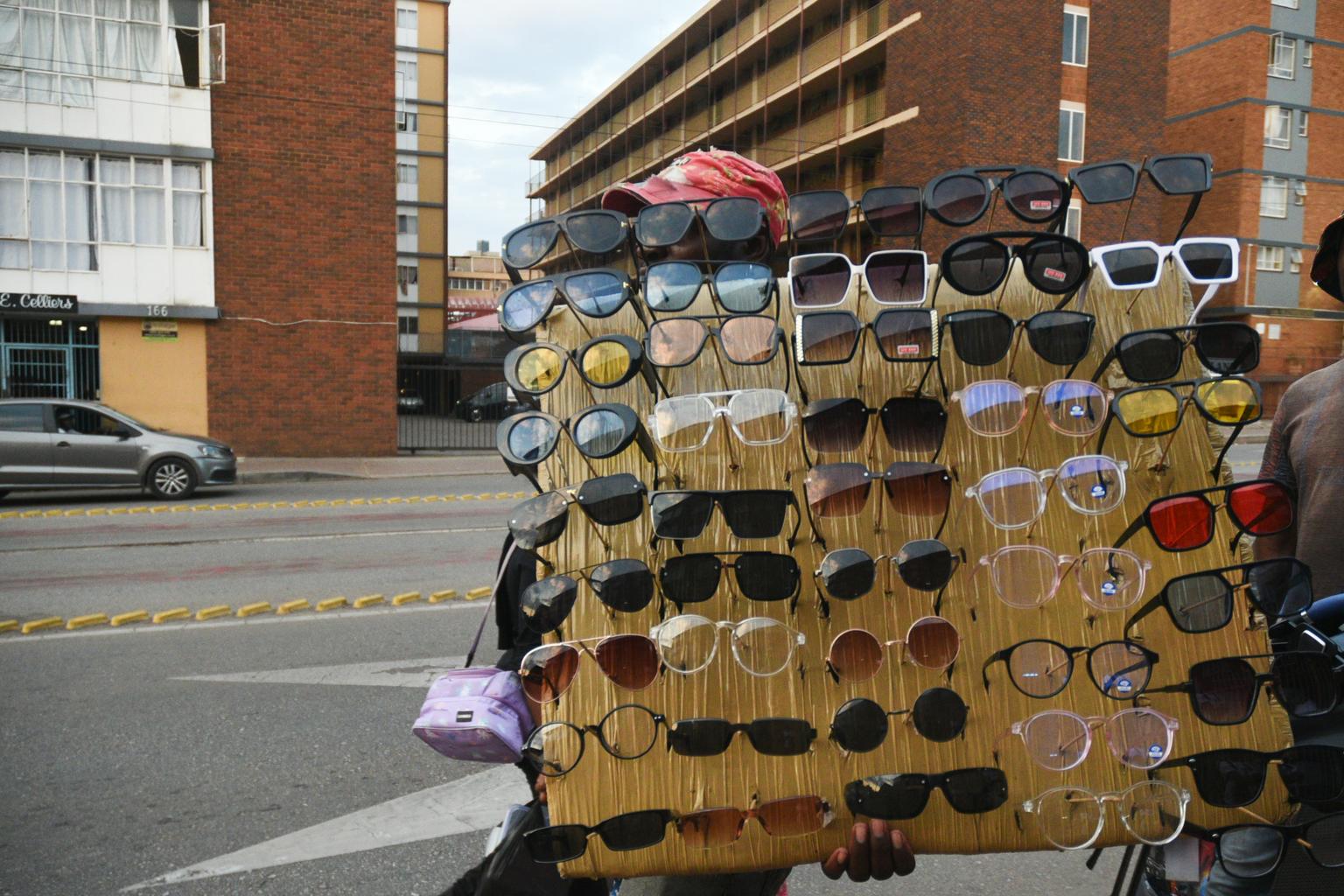 Framed Views: Portable Eyewear Display  A street vendor holds up a dense board lined with dozens of sunglasses and frames against the backdrop of brick apartment blocks.