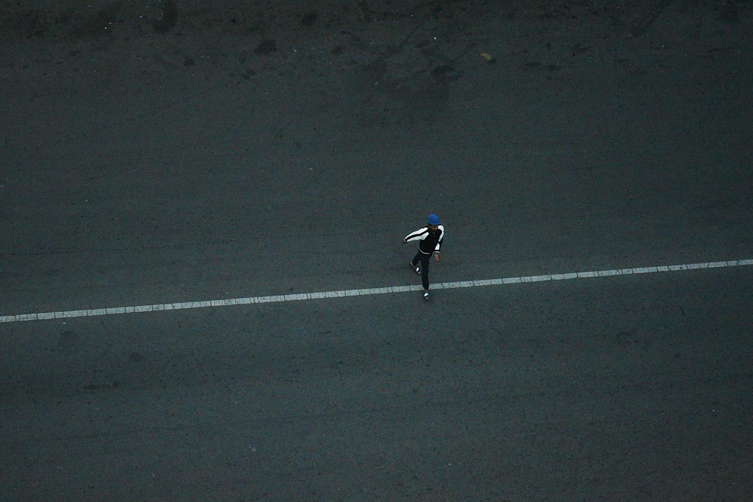 Solitary Crossing: Top-Down Pedestrian: A high-angle vertical shot isolates a solitary figure wearing a blue cap as they step directly across a painted white road line on dark asphalt.
