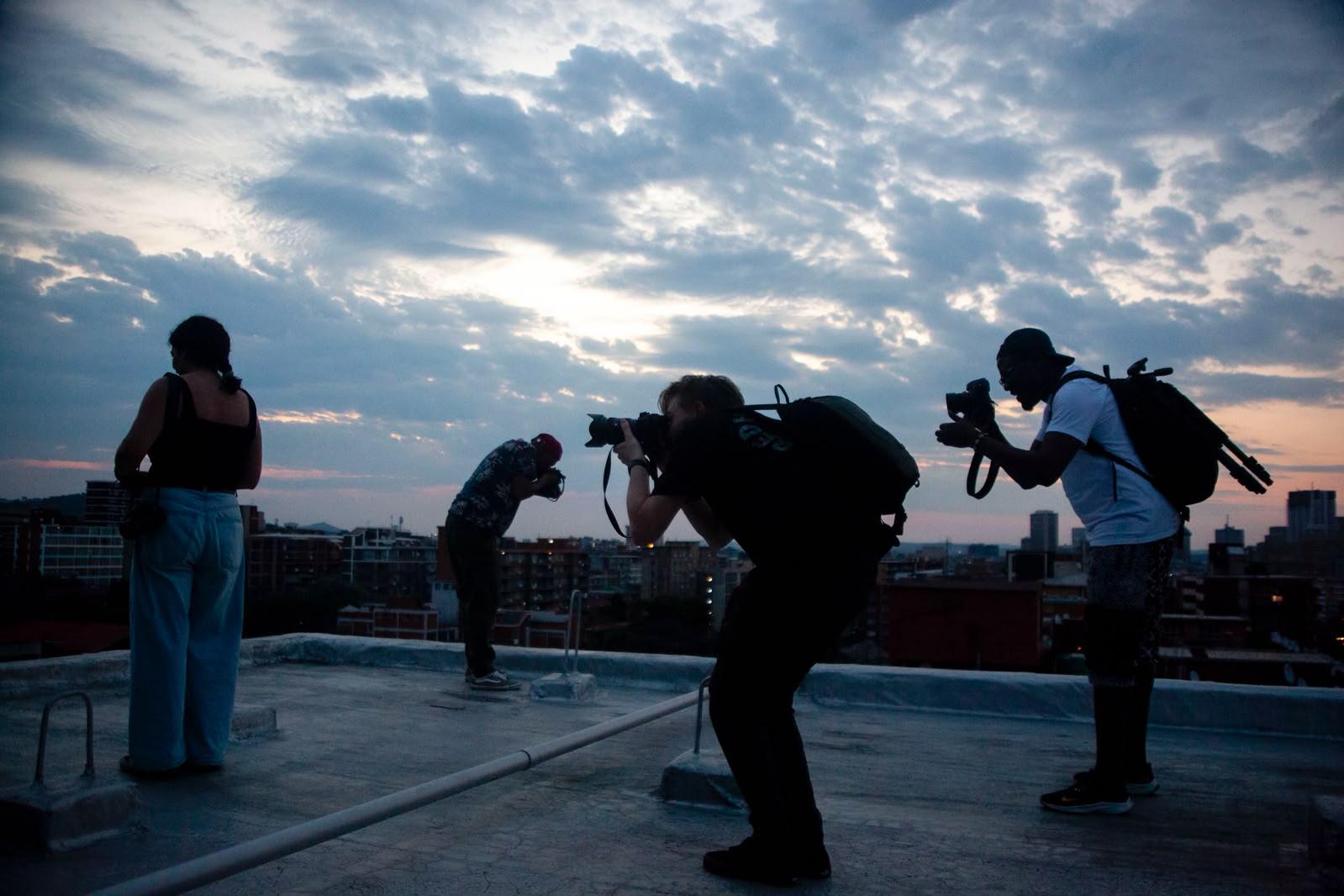 Silhouettes of four people on a rooftop at dusk, three of them bent over their cameras, against a cloudy sky with the city below.