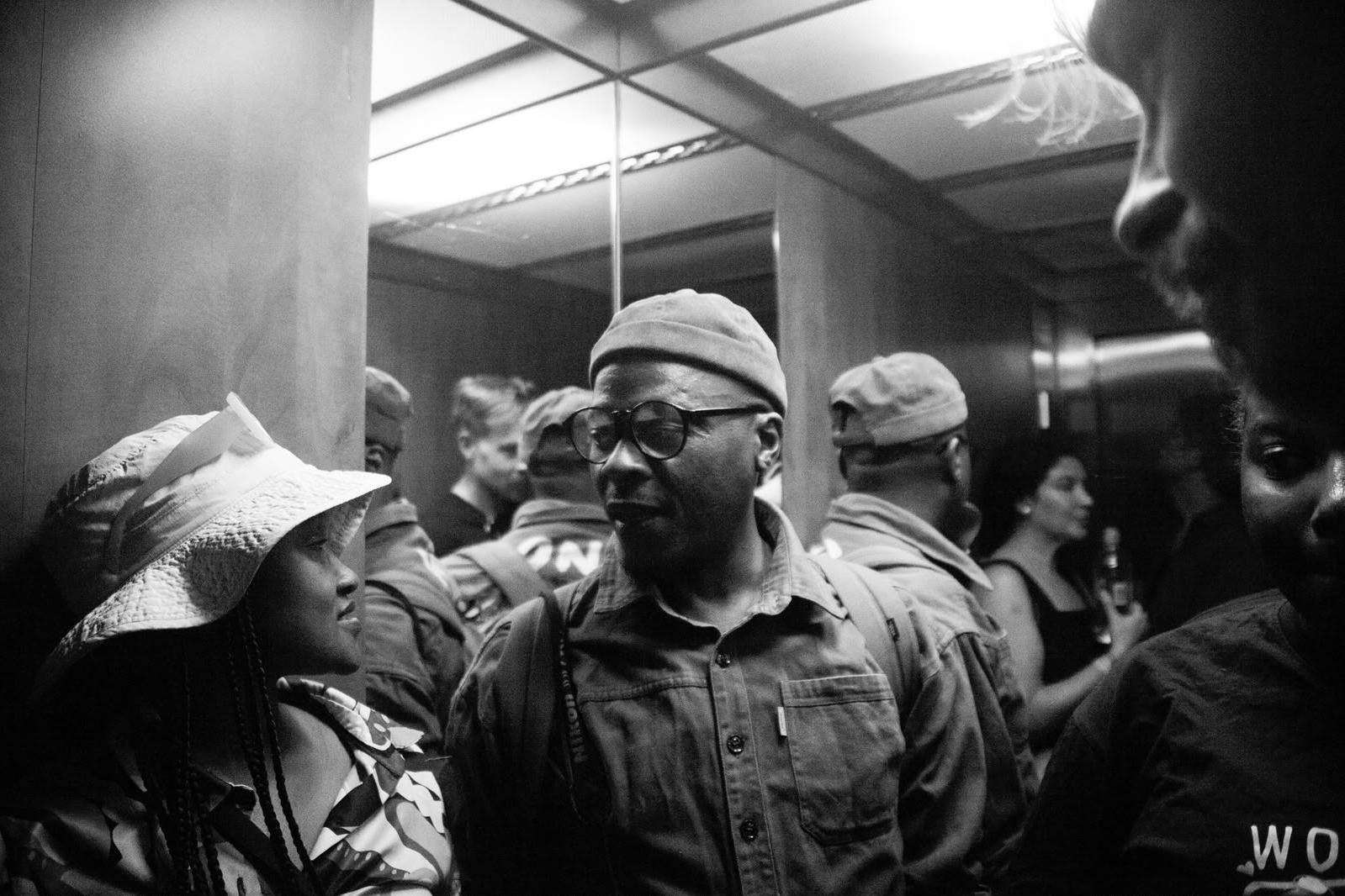 Black and white photograph of a crowded mirrored lift, with a man in glasses and a beanie in the centre talking to a woman in a white bucket hat.
