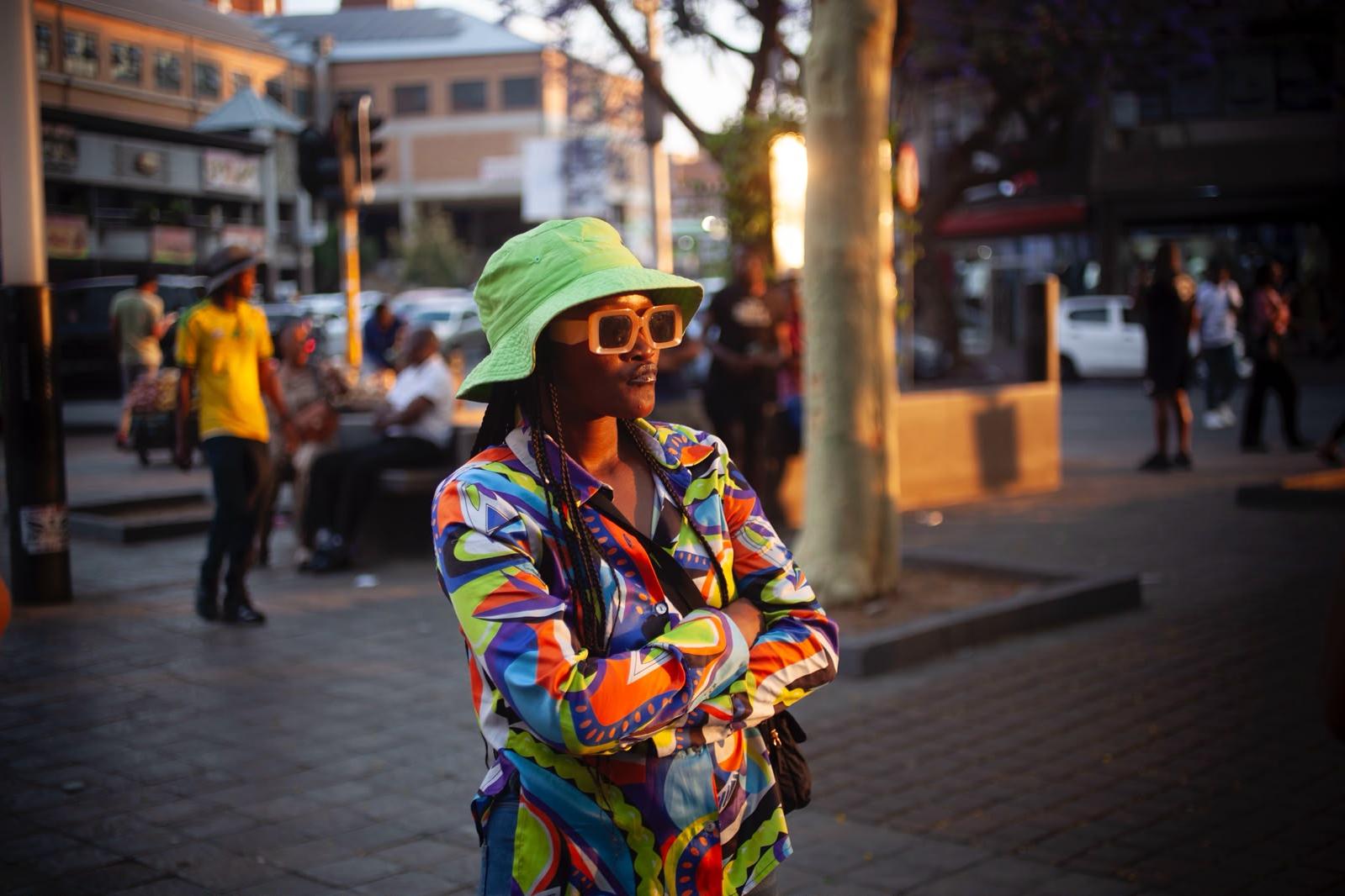 A woman in a lime green bucket hat, oversized cream sunglasses and a bright patterned shirt stands with folded arms on a busy pavement, lit by low golden sun.