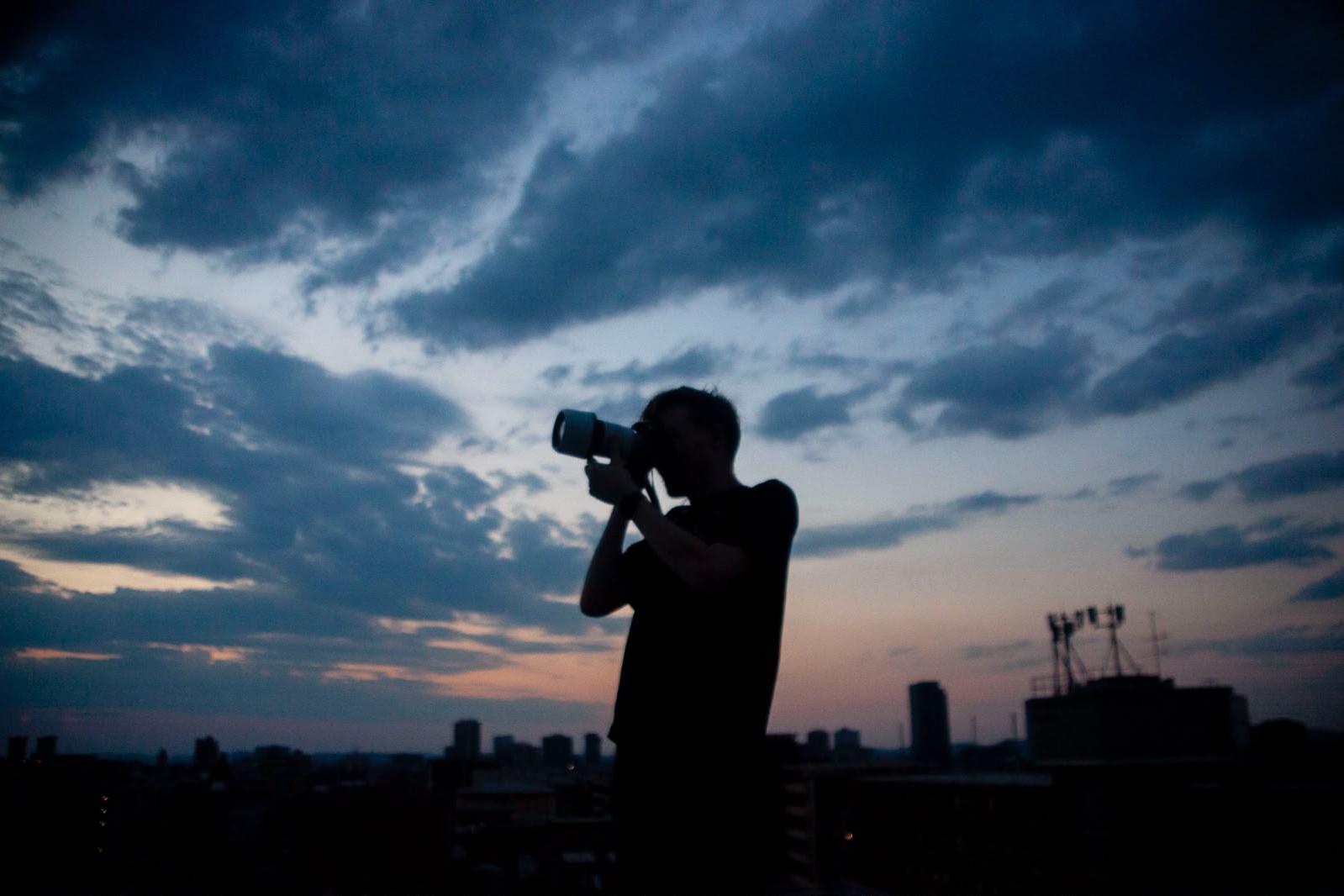 The silhouette of a man raising a camera with a long lens against a deep blue evening sky, with a dark city skyline and antenna towers behind him.