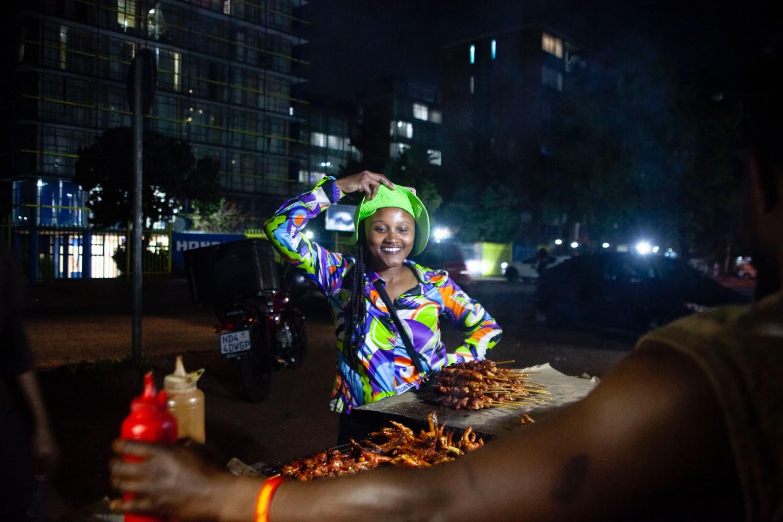 A smiling woman in a lime green bucket hat and patterned shirt stands behind a street braai with chicken skewers and chicken feet at night, while a vendor's arm holds a sauce bottle in the foreground.