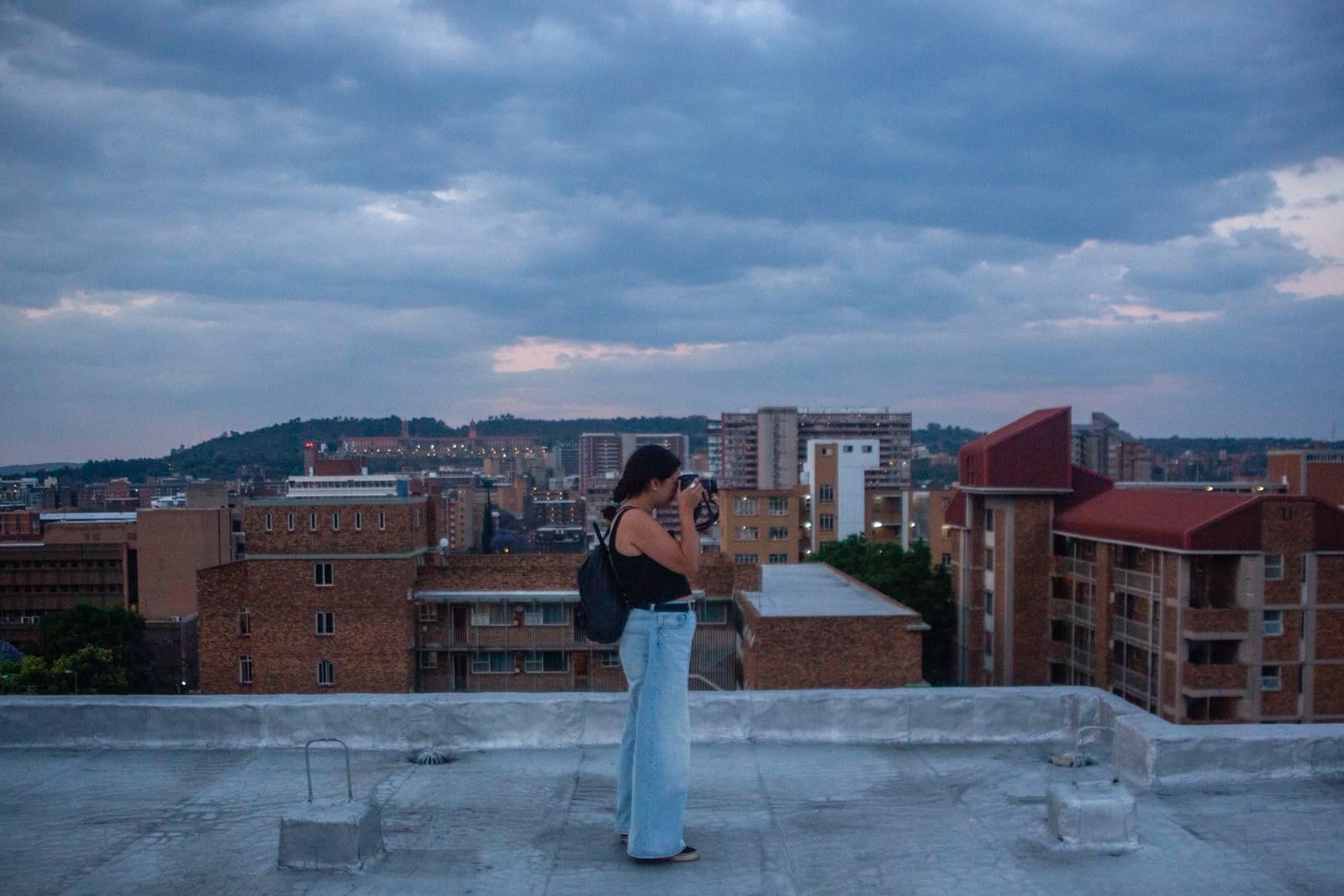 A young woman in wide light blue jeans and a black top photographs the city from a flat rooftop, with red brick apartment blocks and a lit hilltop building under a heavy grey sky.