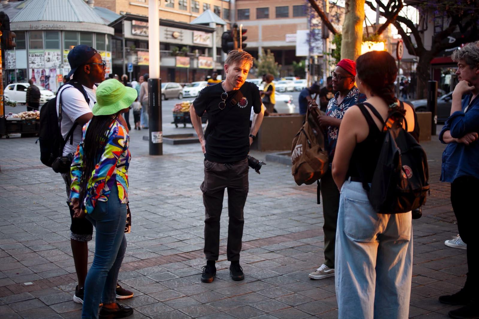 A man in a black T-shirt talks to a circle of photographers with backpacks and cameras on a paved Sunnyside street corner in late afternoon light.