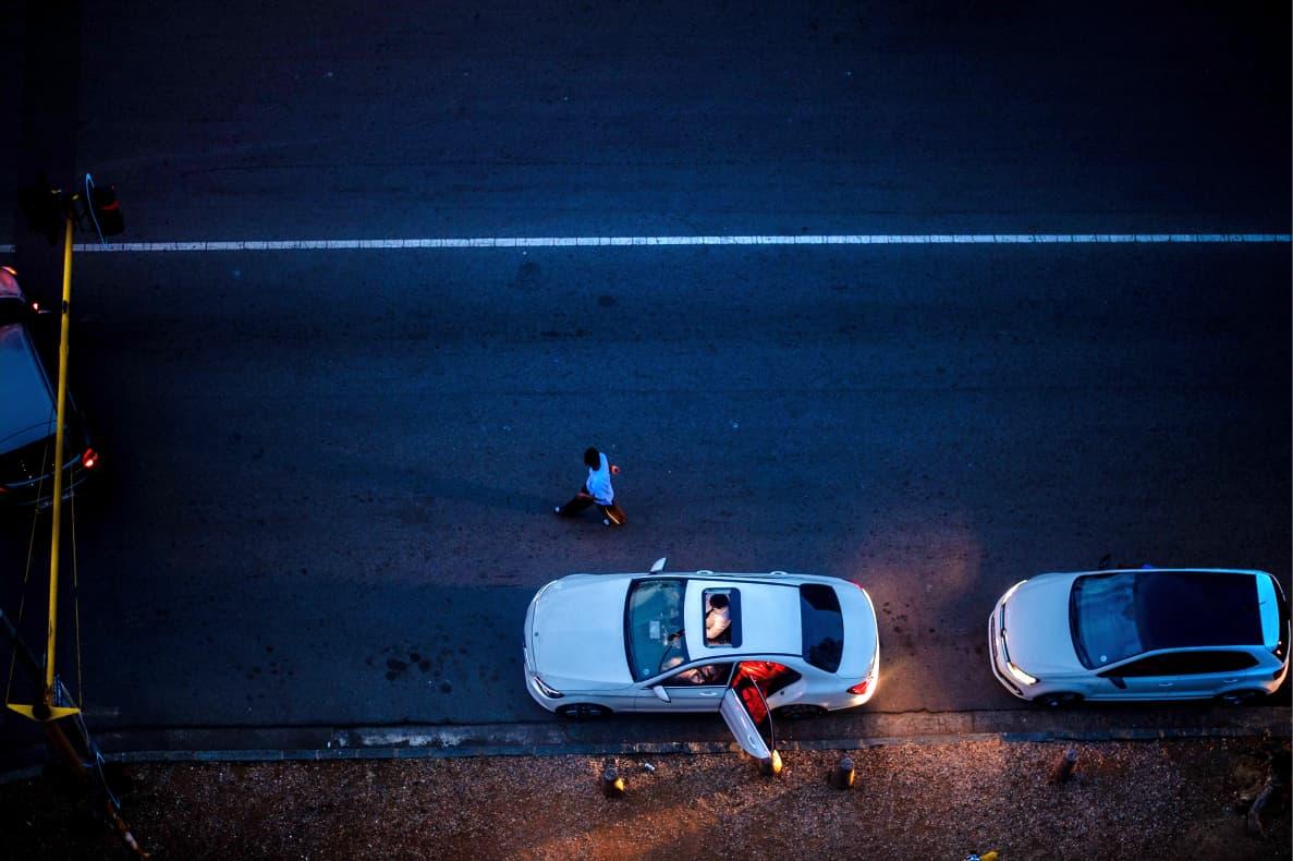 Top-Down Motion: Street Overhead: A high-angle bird’s-eye view captures a pedestrian navigating the dark asphalt between parked vehicles illuminated by warm headlights.