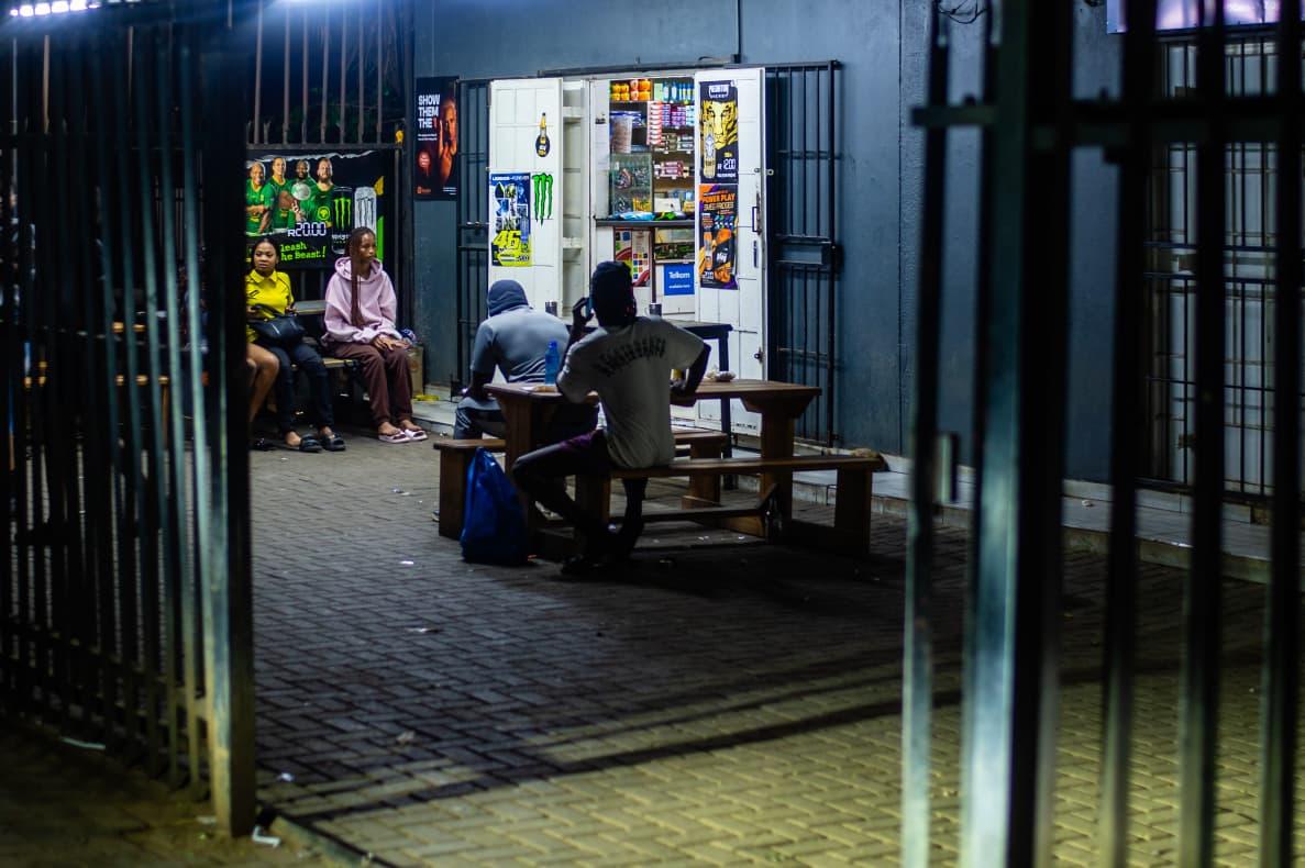 Nighttime Gathering: Spaza & Sidewalk Patrons relax on wooden benches outside a brightly lit kiosk, framed by dark foreground security gates and colorful promotional posters.