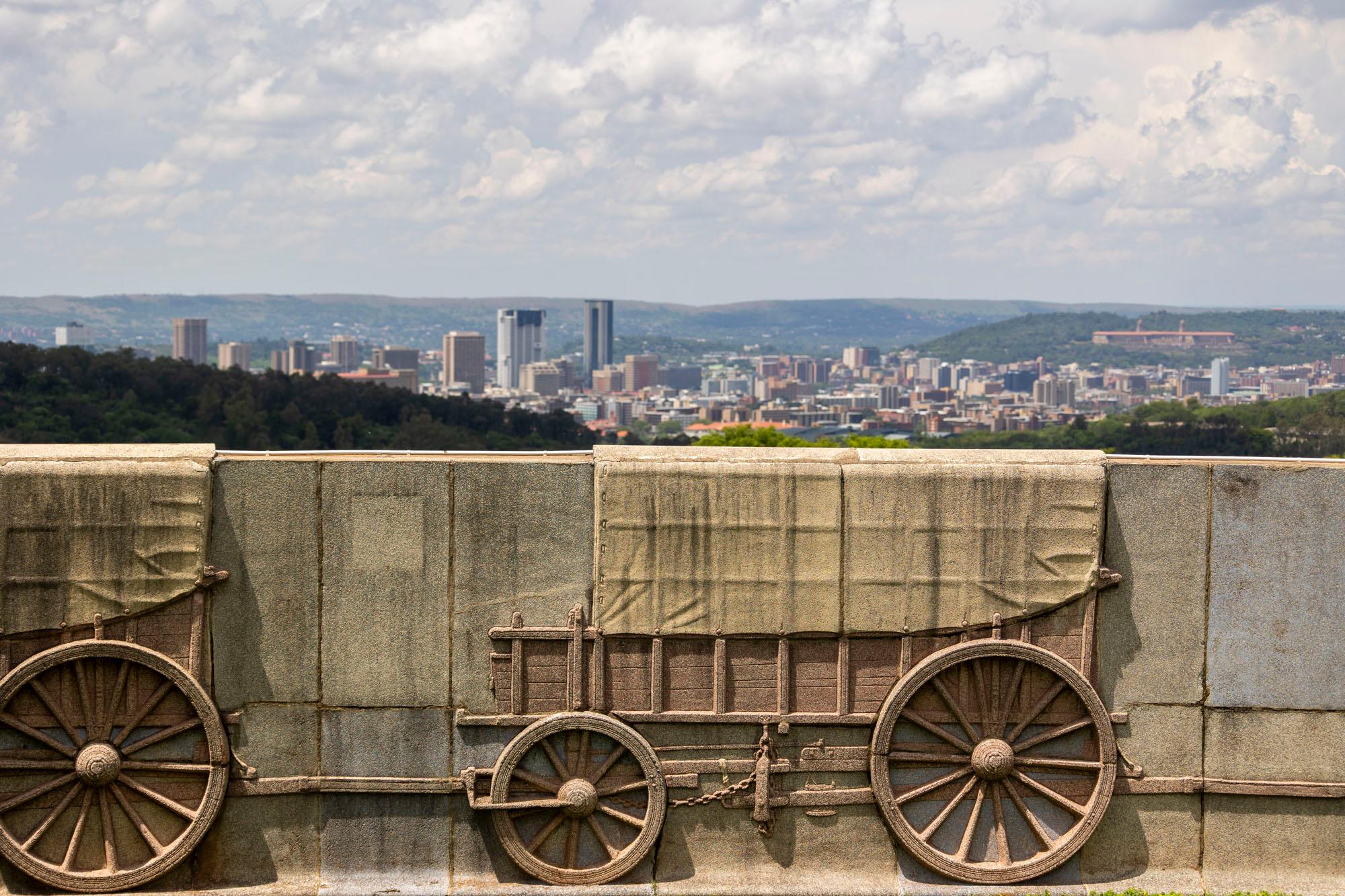 A carved stone relief of ox wagons on the Voortrekker Monument's outer wall, with the Pretoria skyline and hills visible behind it.