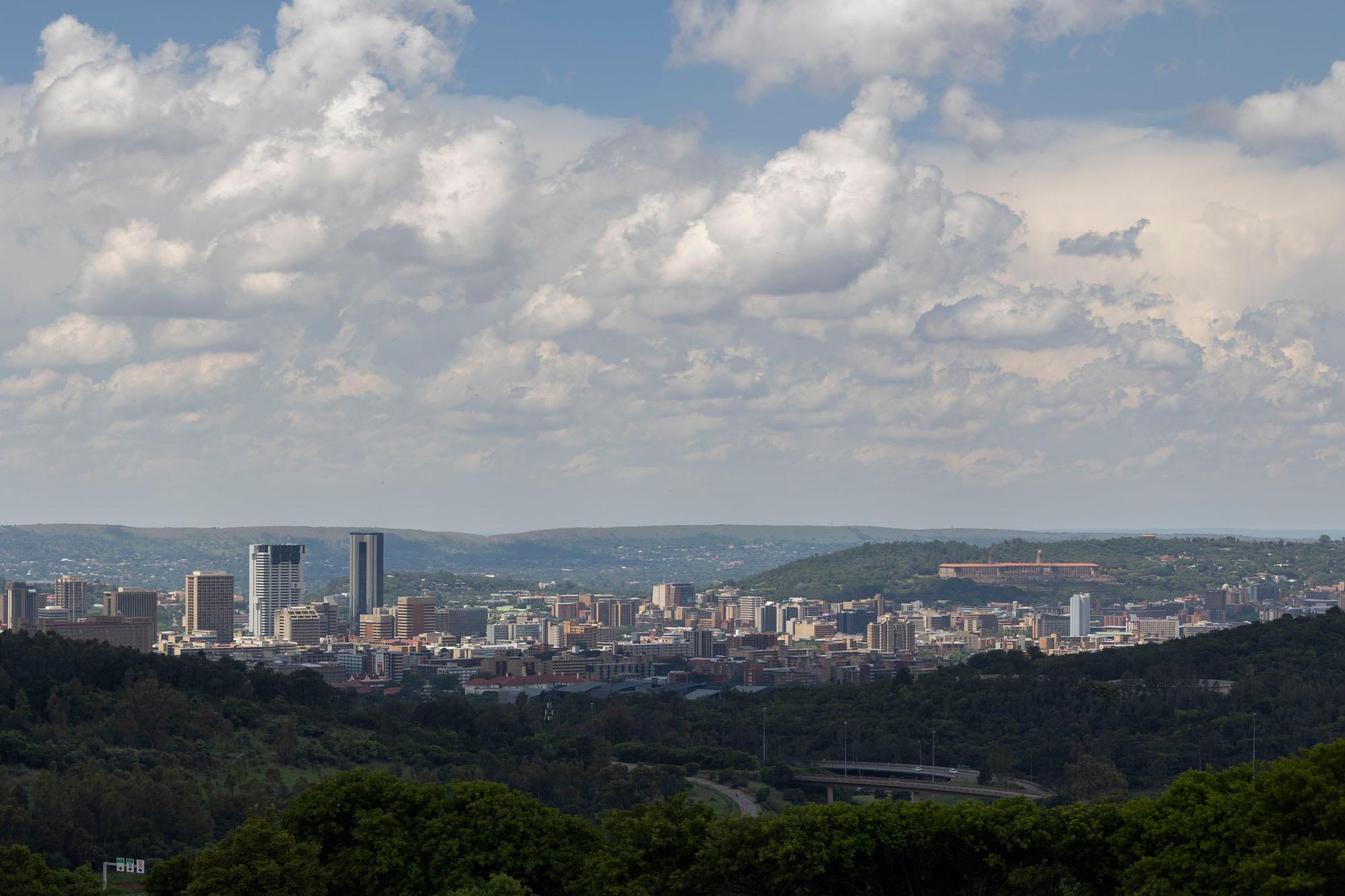 Wide view of the Pretoria city centre skyline from a wooded ridge, with the Union Buildings on the hillside to the right and cumulus cloud above.