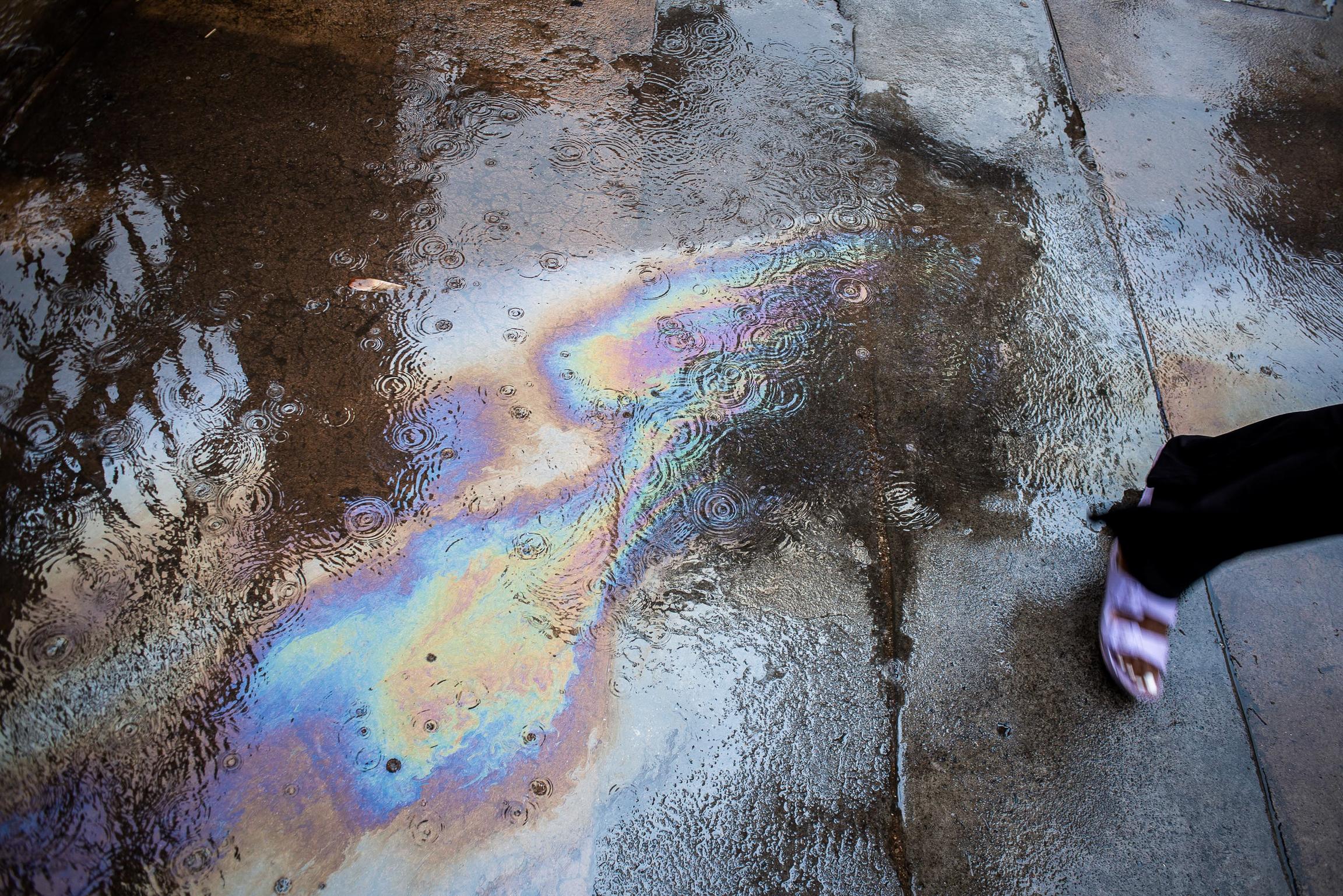 An oil slick spreads bands of colour across a puddle on tar, the edge of a foot at the frame edge.