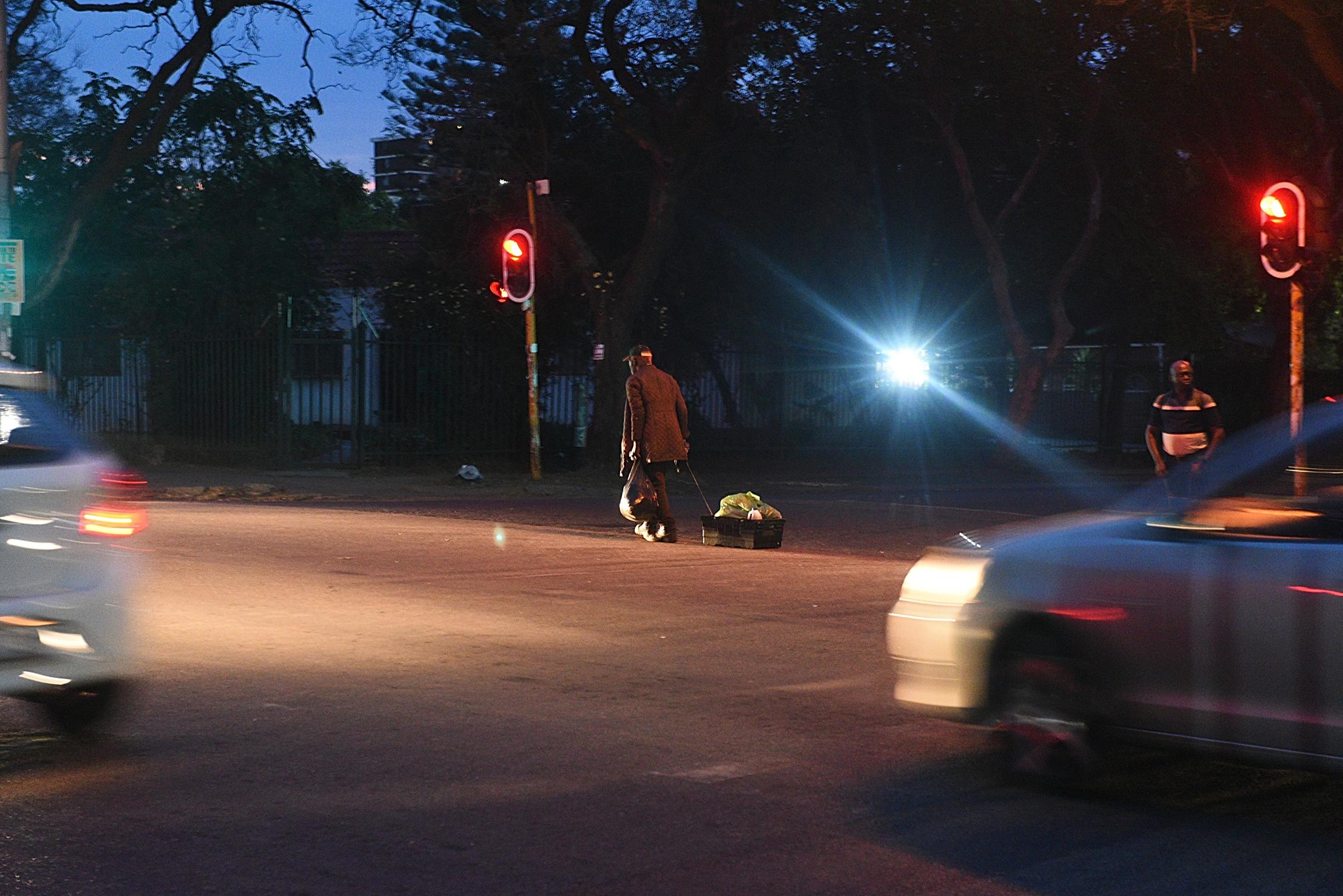Intersections at Twilight: Crossing the Street:  Red traffic signals and glaring car headlights slice through the dusk as a pedestrian pulls a crate across a quiet intersection. 