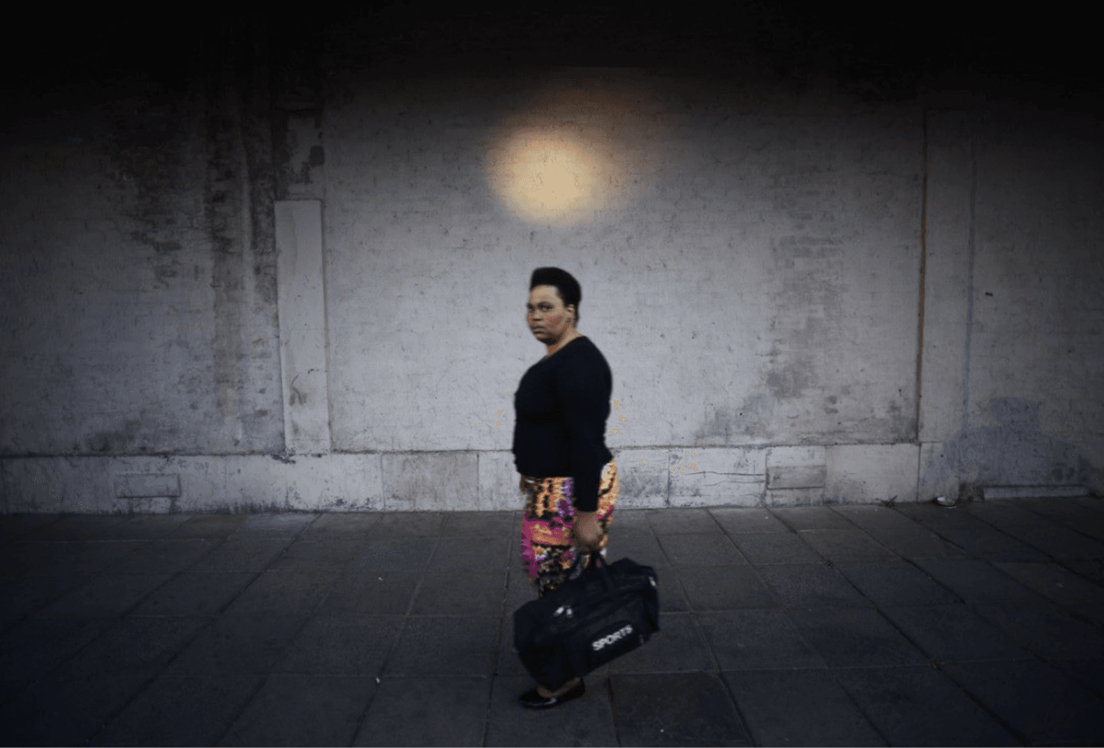 A woman in a black top and patterned skirt walks along a pavement carrying a sports bag, beneath a round pool of light on a dark wall.