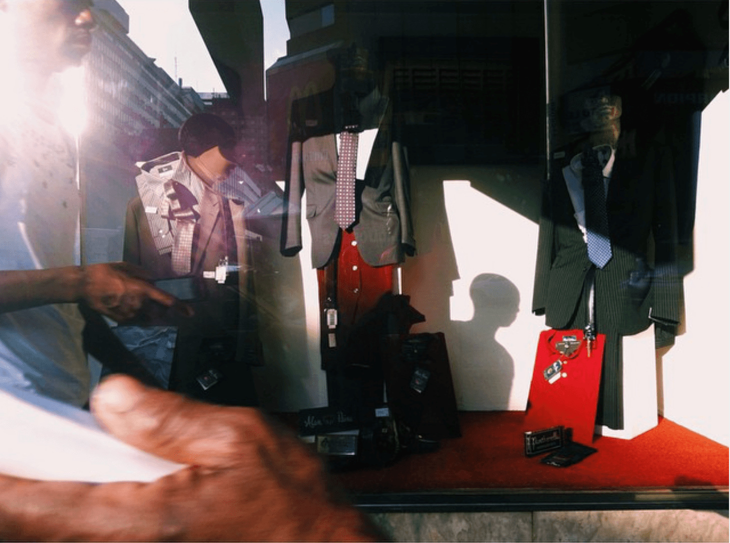 A menswear display window of suits and ties on hangers, a passer-by reflected in the glass and a head-shaped shadow on the back wall.