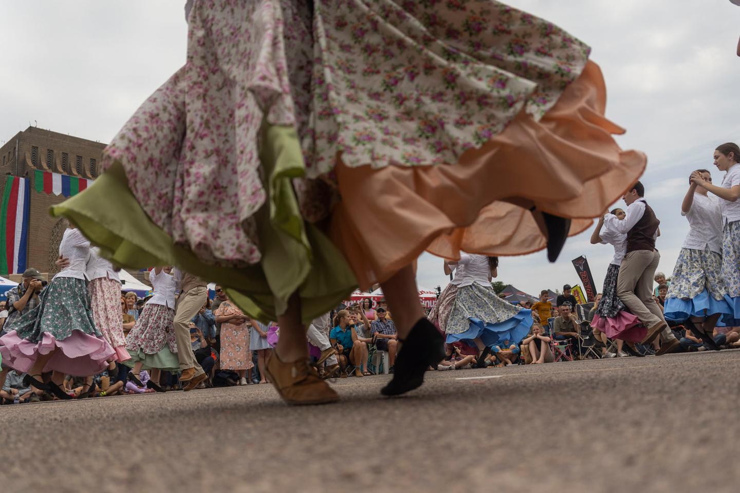 Low-angle view of folk dancers in long floral skirts and boots turning on tarmac, with a seated crowd and festival tents behind.