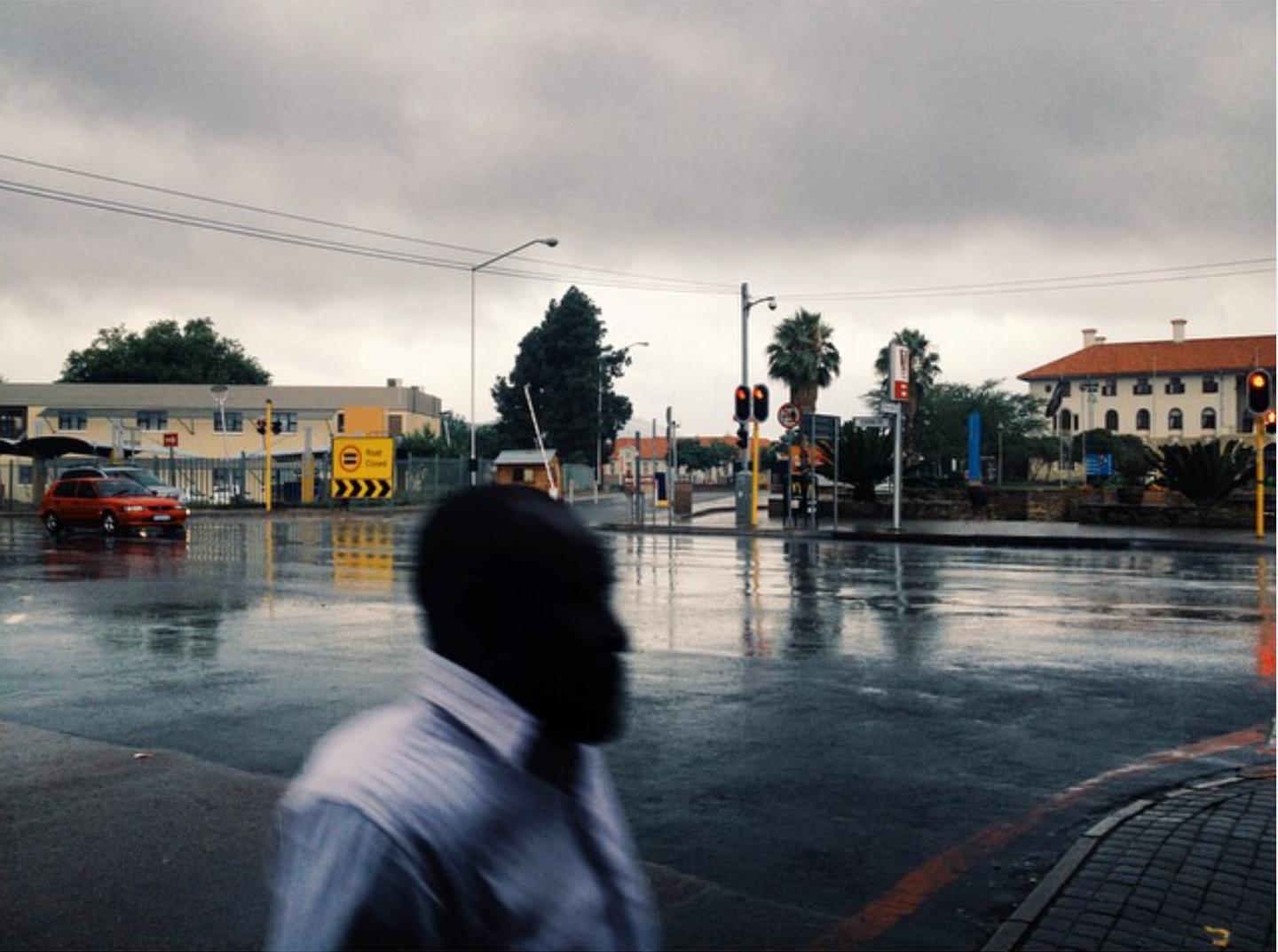 A man in a white shirt walks through a rain-soaked intersection at dusk, his head a dark silhouette against wet tar.