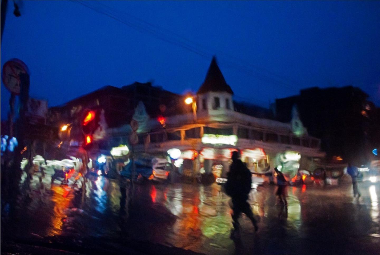Blurred blue-hour street scene in rain, taxi and shop lights reflecting on a flooded road, a dark figure walking across the centre.