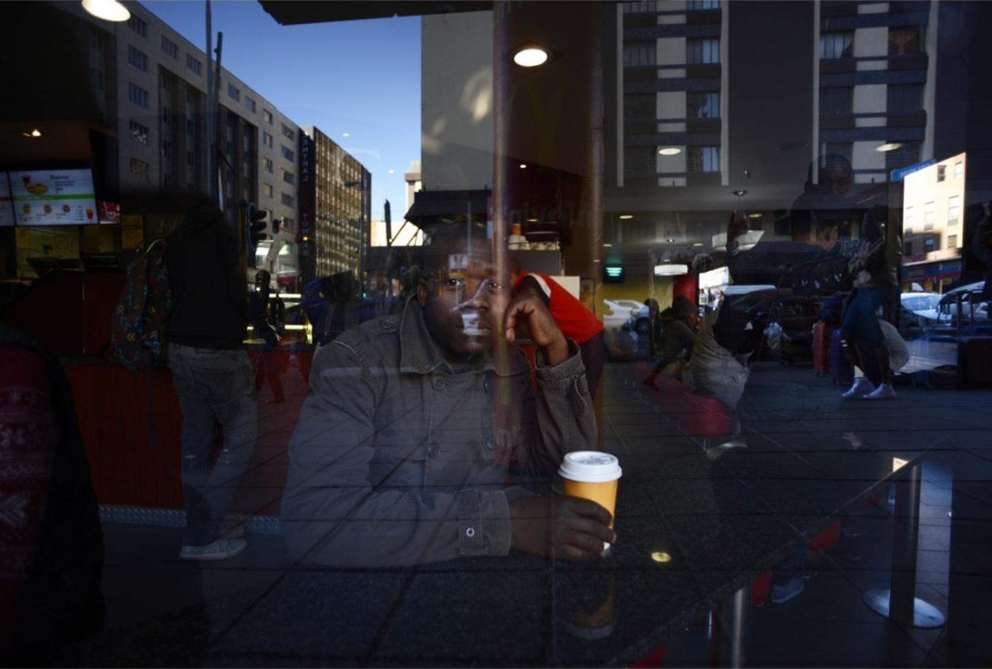A man sits with a takeaway coffee cup behind a window, the reflected street and buildings overlaying his face and jacket.