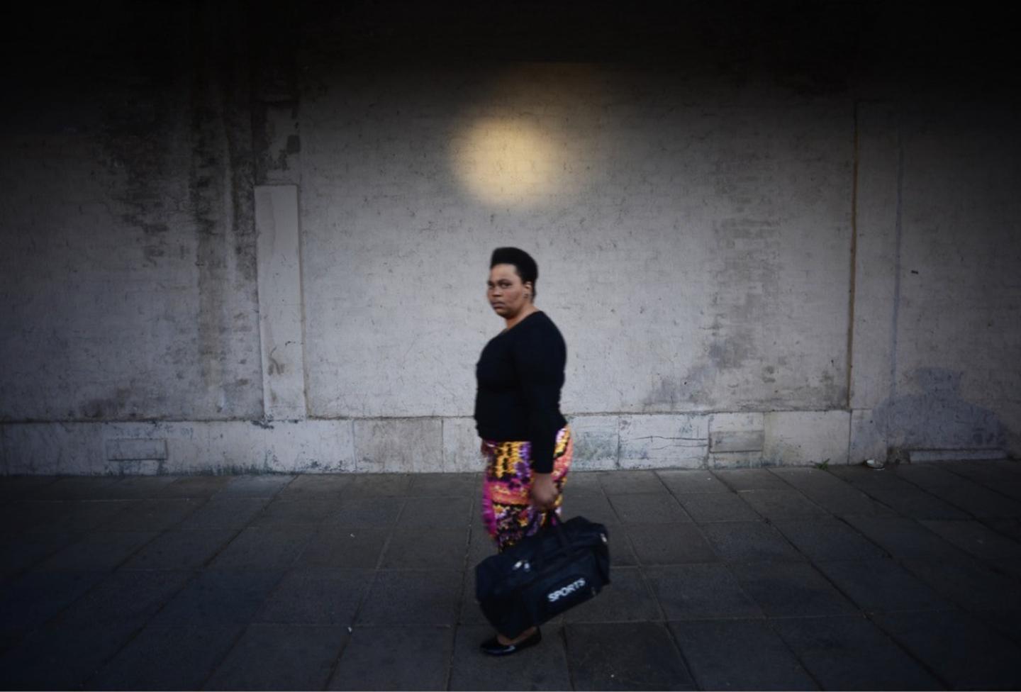 A woman in a black top and patterned skirt walks along a pavement carrying a sports bag, beneath a round pool of light on a dark wall.