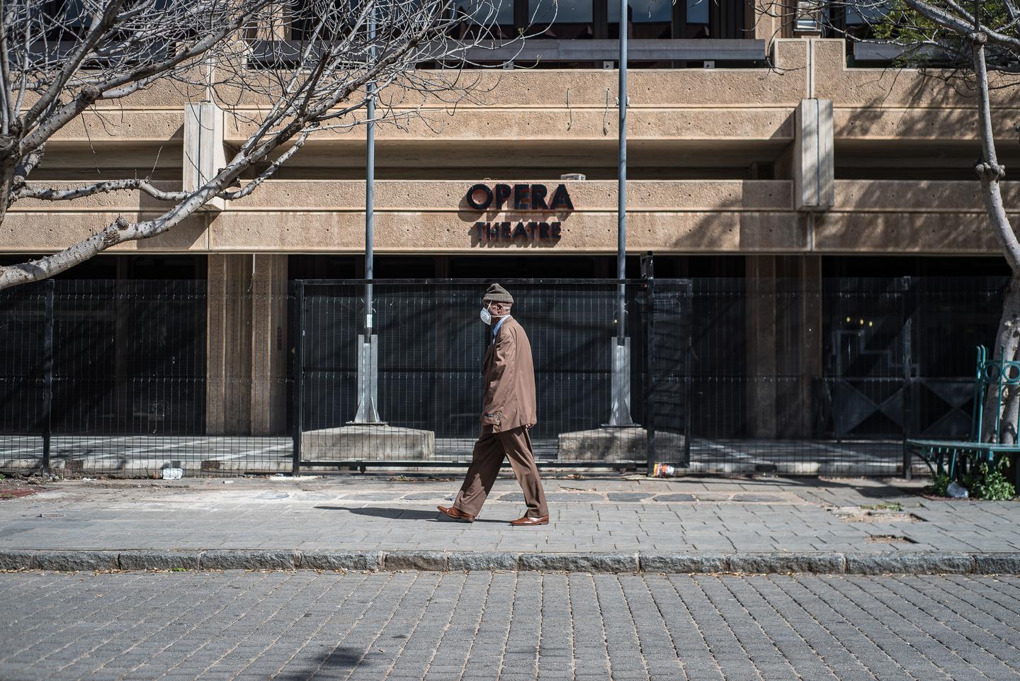A man walks along an empty pavement past a building signed Opera, one pigeon in the air.