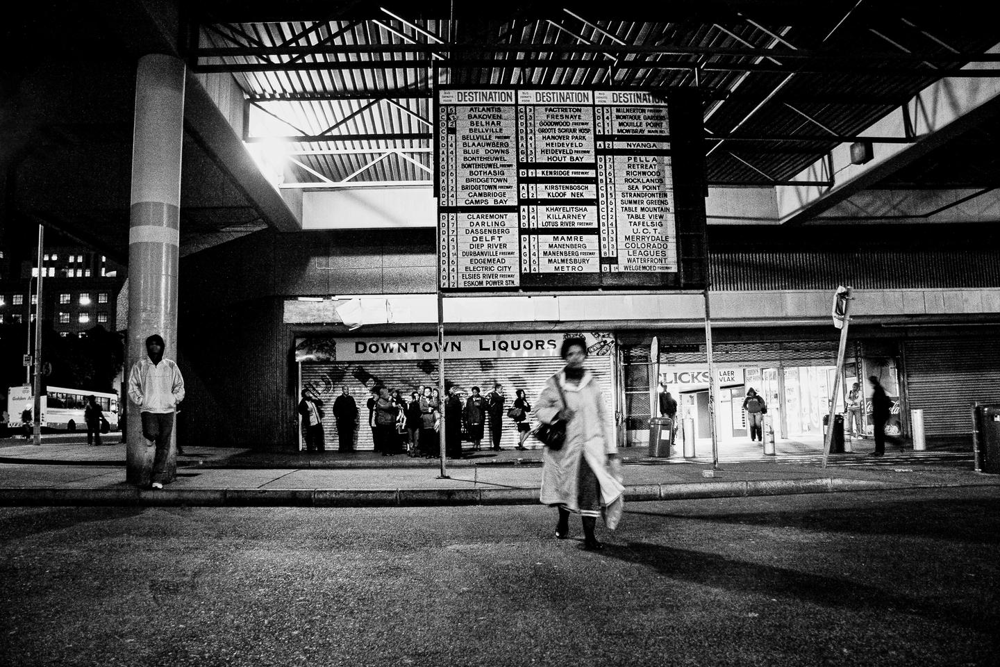Central Cape Town bus terminus and a large bus destination board listing dozens of place names, with people waiting beneath it outside shuttered shops.