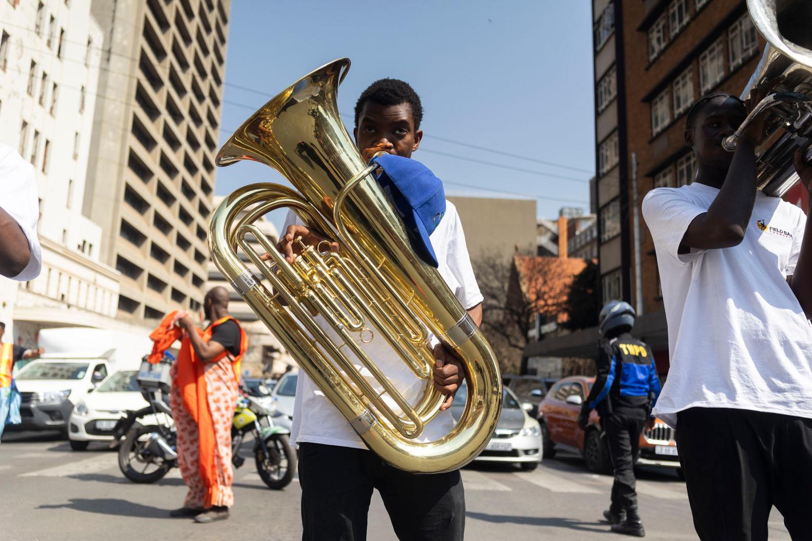 A young man plays a large brass tuba while marching in a city street, another player beside him, with cars, a traffic officer and high-rise buildings around them.