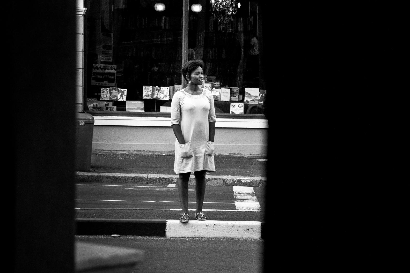 A woman in a pale dress standing on a narrow traffic island in front of a bookshop window, framed by dark vertical edges.
