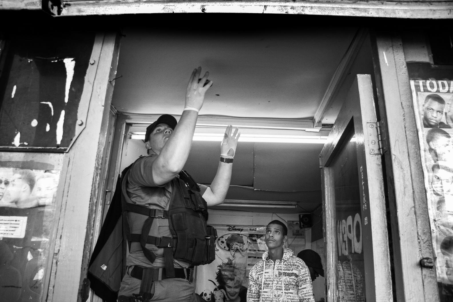Photograph taken from below of a police officer in a tactical vest with both hands raised in a shop doorway, two men standing inside behind him.
