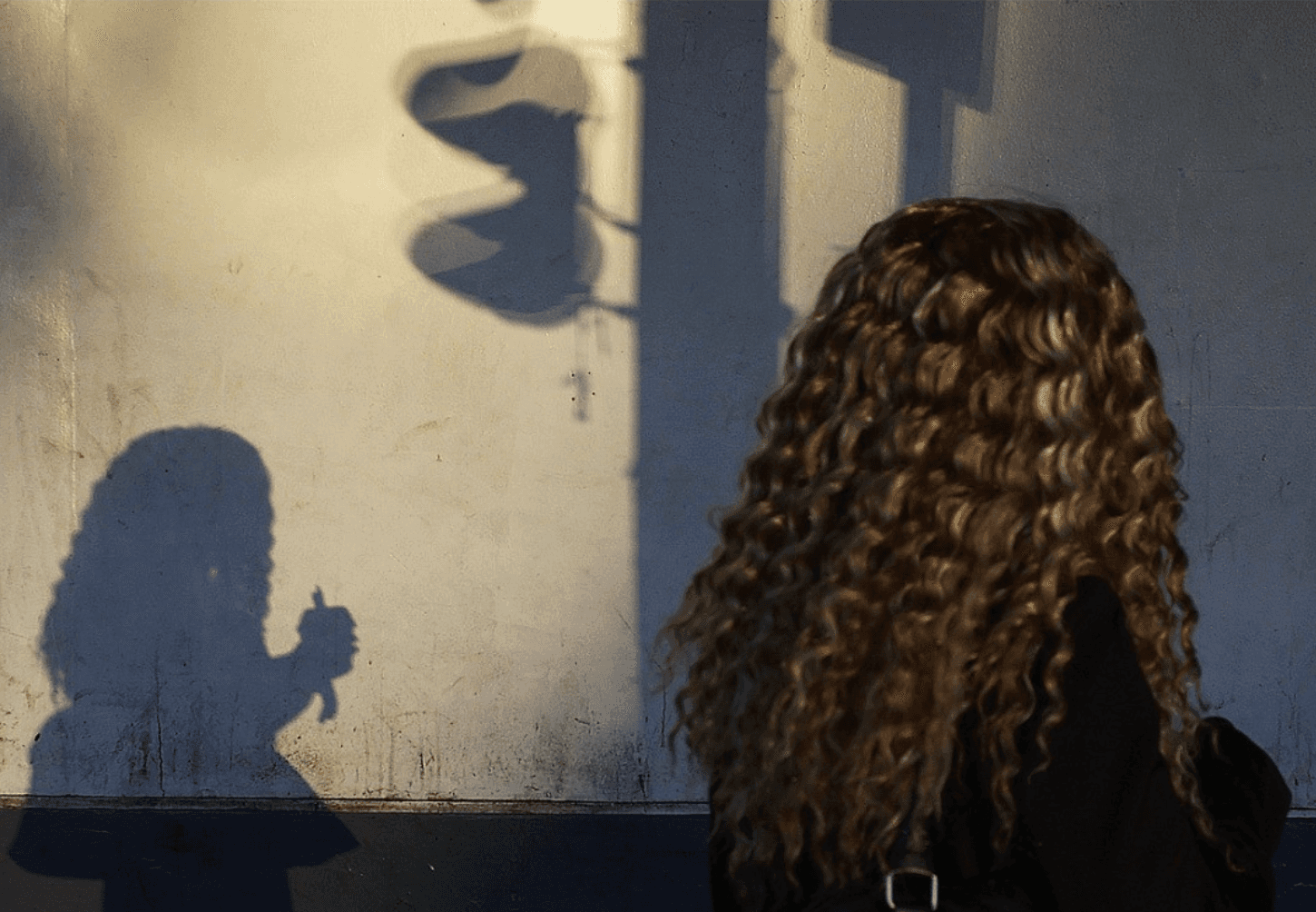 A woman with long curly hair, seen from behind, faces a sunlit wall where her shadow holding a cup falls beside a traffic-light shadow.