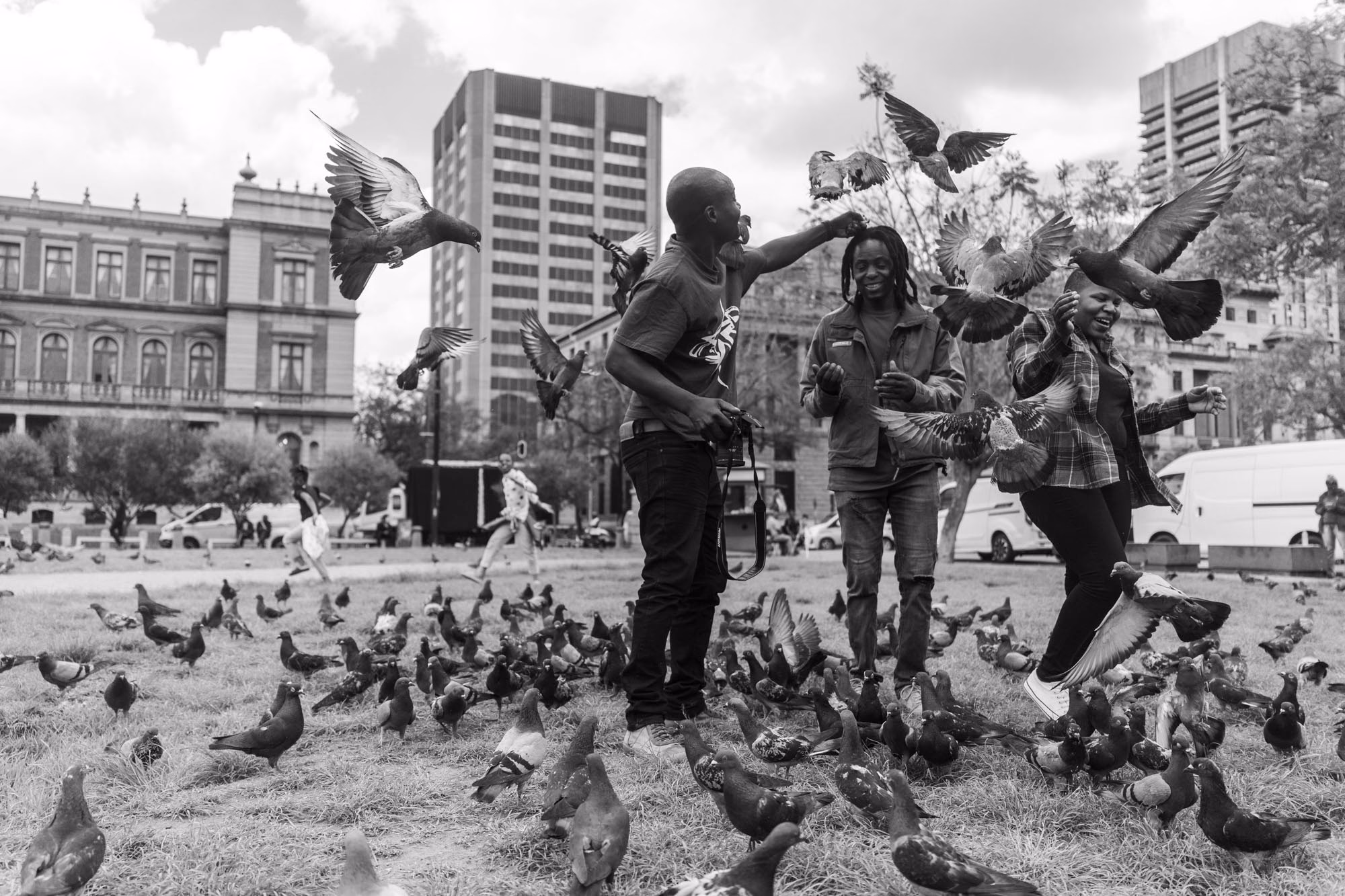 Black and white photograph of three people on a city square lawn surrounded by hundreds of pigeons, several birds in flight around their outstretched hands.