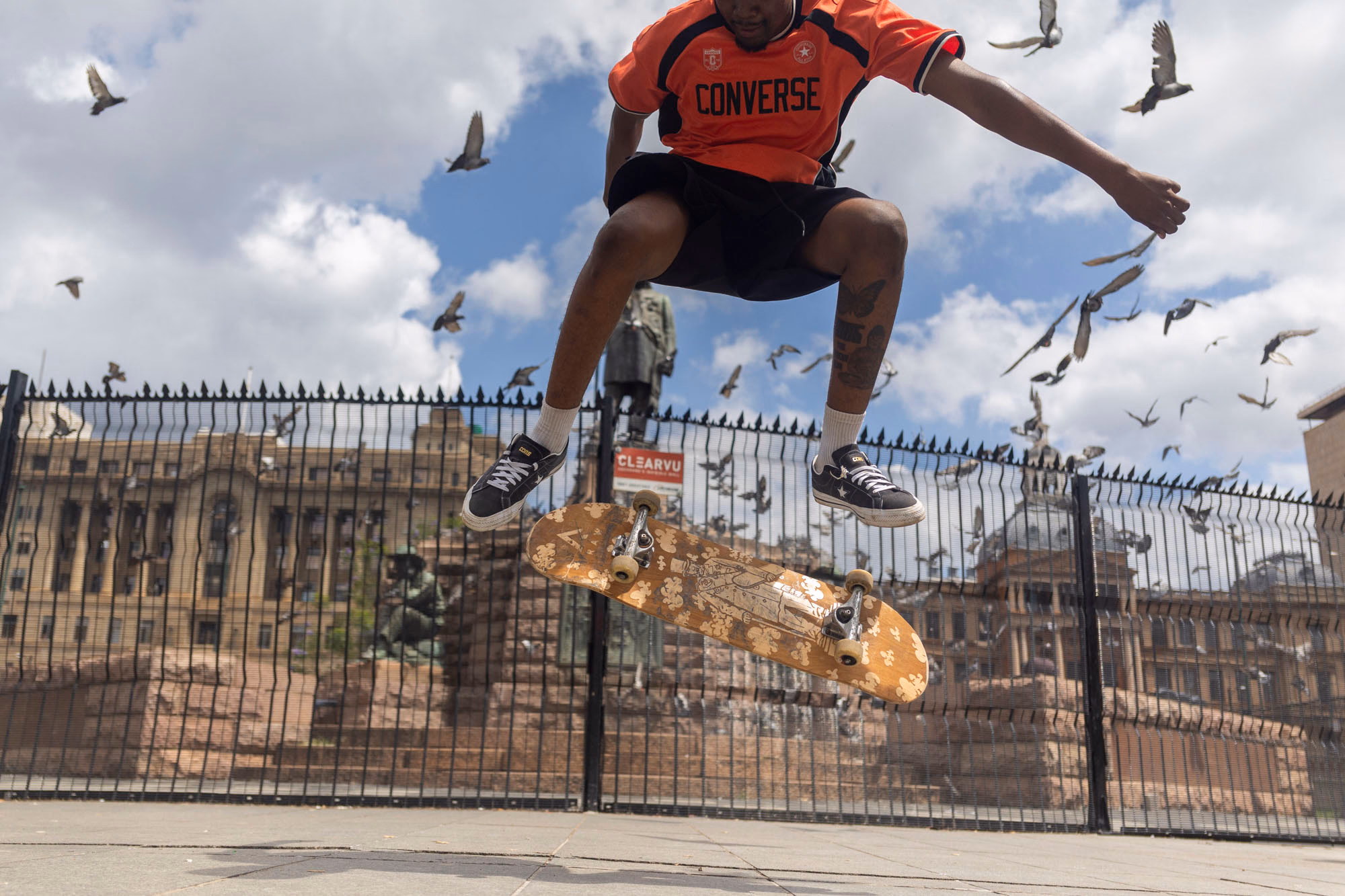 A skater in a red Converse shirt in mid-air above his board, pigeons scattering around him, with a palisade fence and a sandstone government building behind.