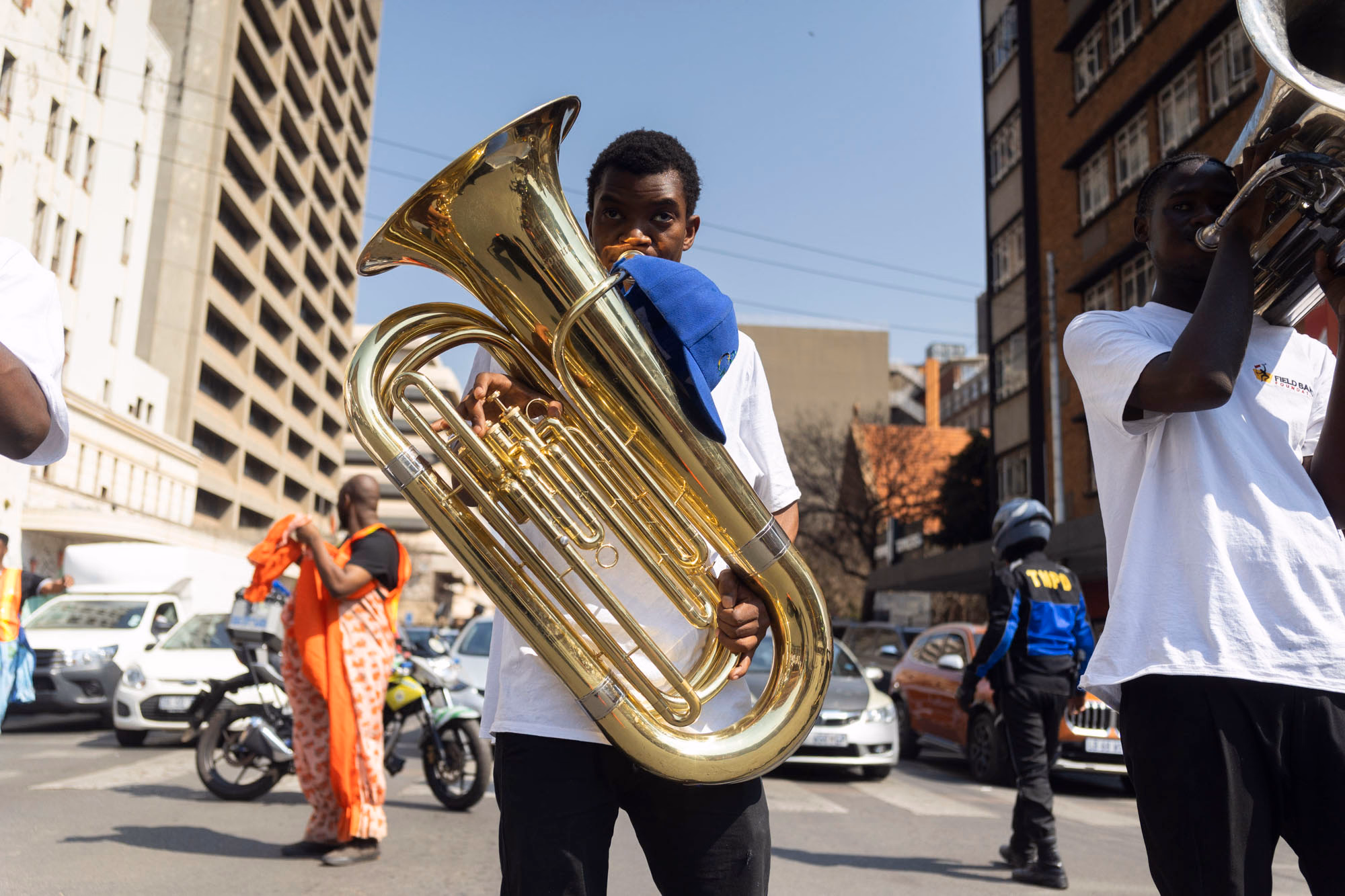 A young man plays a large brass tuba while marching in a city street, another player beside him, with cars, a traffic officer and high-rise buildings around them.