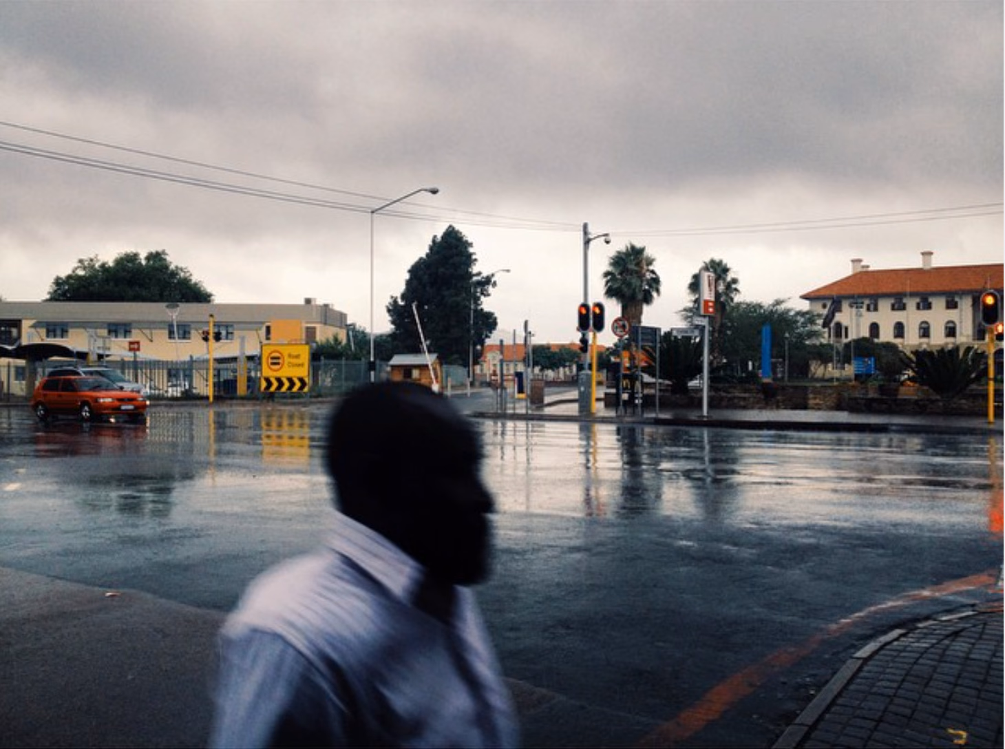 A man in a white shirt walks through a rain-soaked intersection at dusk, his head a dark silhouette against wet tar.