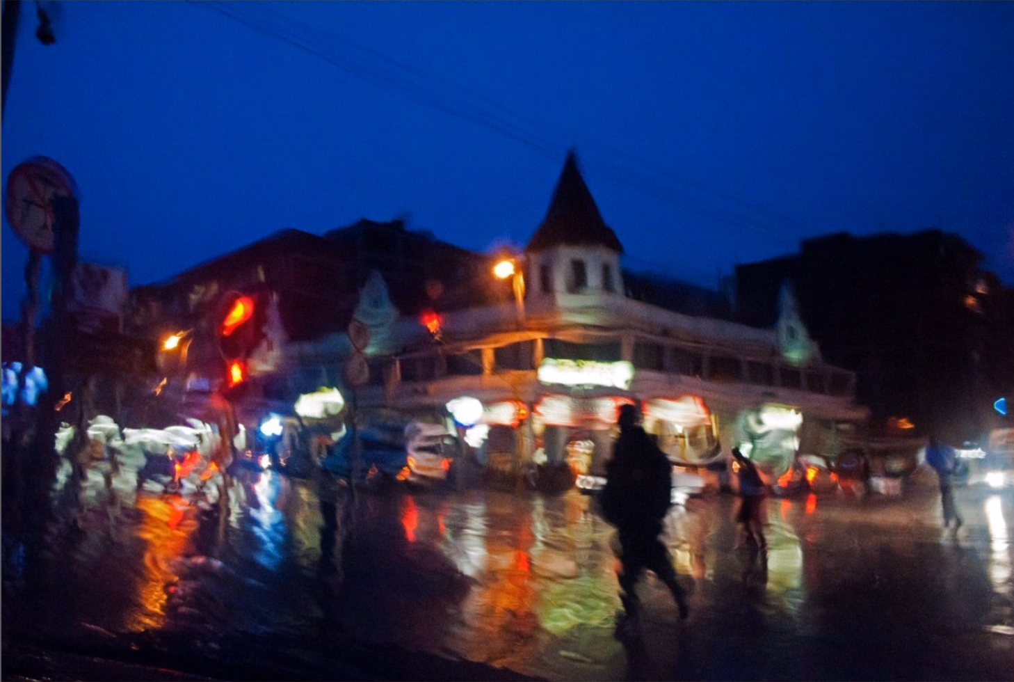 Blurred blue-hour street scene in rain, taxi and shop lights reflecting on a flooded road, a dark figure walking across the centre.
