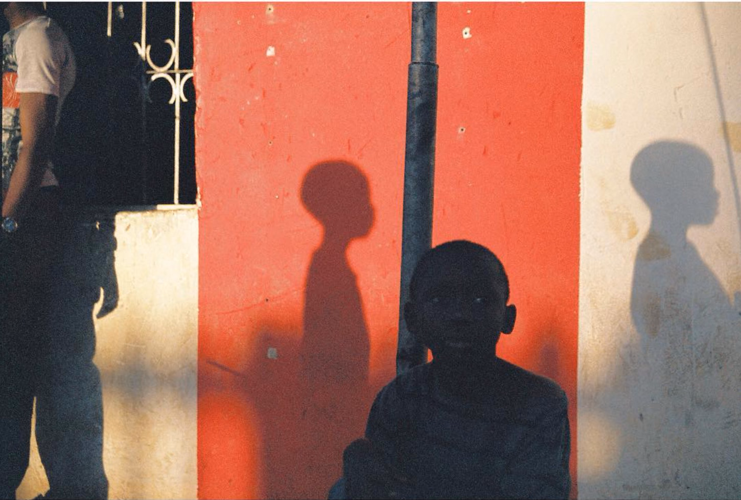 A young boy in silhouette sits against a red and white painted wall, with two head-and-shoulder shadows falling on either side of him.