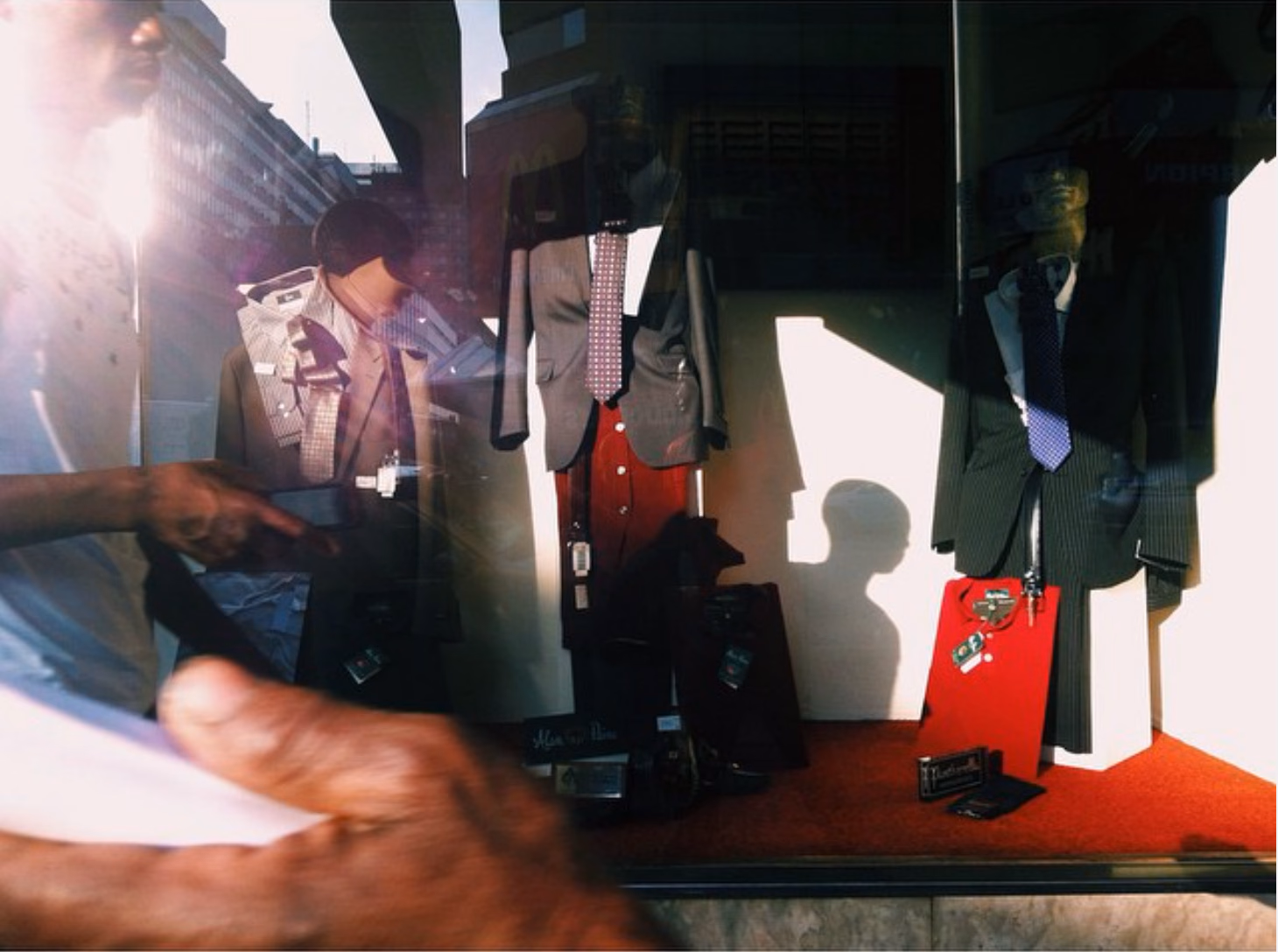 A menswear display window of suits and ties on hangers, a passer-by reflected in the glass and a head-shaped shadow on the back wall.