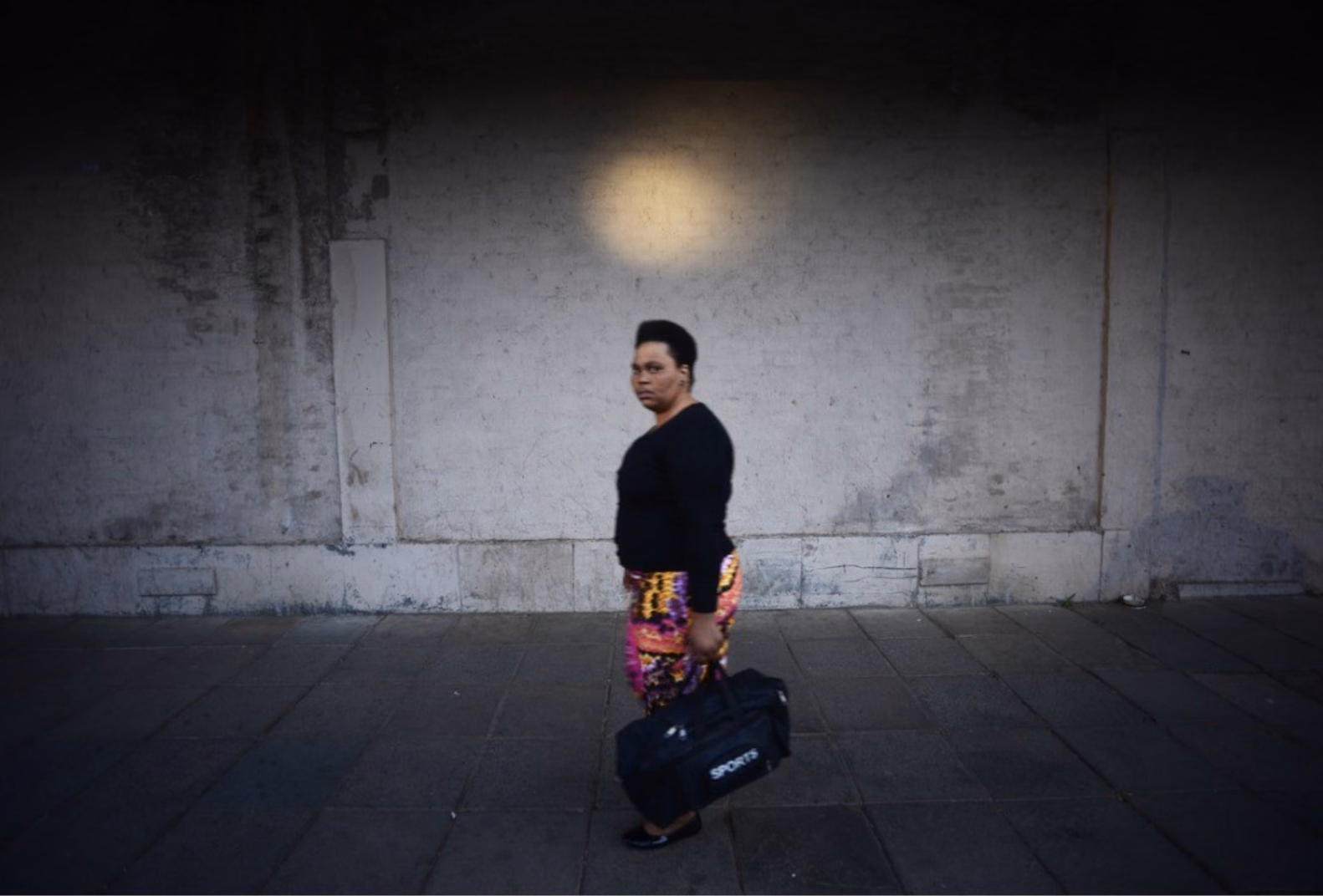 A woman in a black top and patterned skirt walks along a pavement carrying a sports bag, beneath a round pool of light on a dark wall.