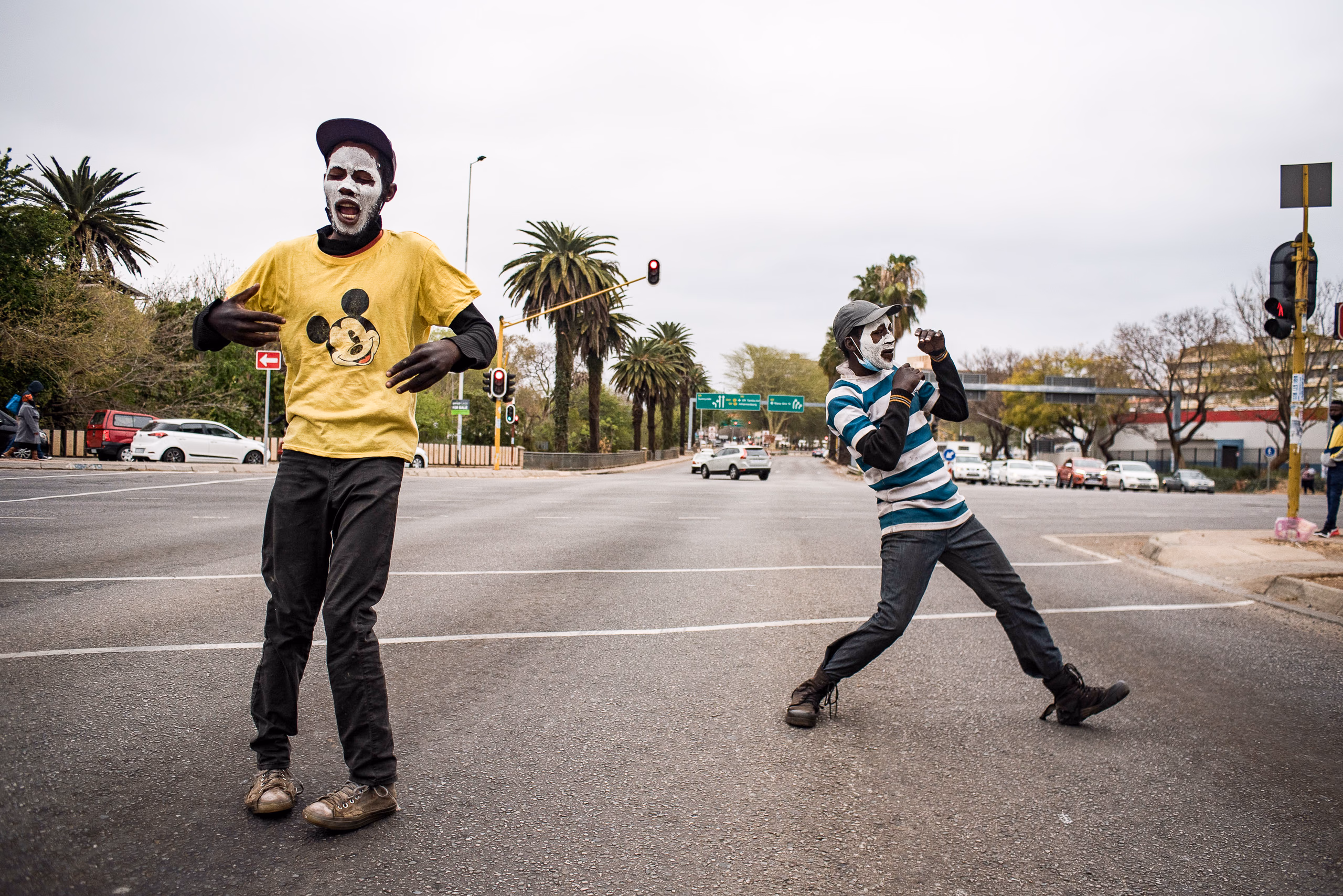 Two men move in the middle of an empty street, one in a yellow shirt, trees and buildings behind.