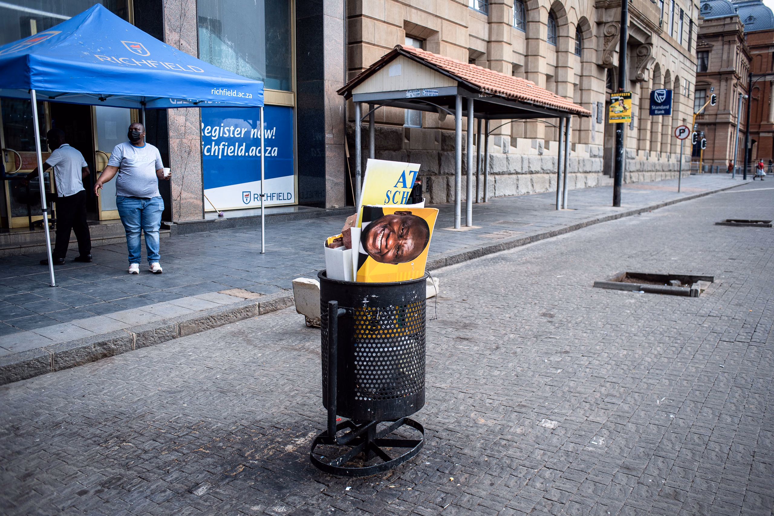 Ramaphosa in the trash bin with a blue gazebo over a street stall on an empty pavement, an older building behind.