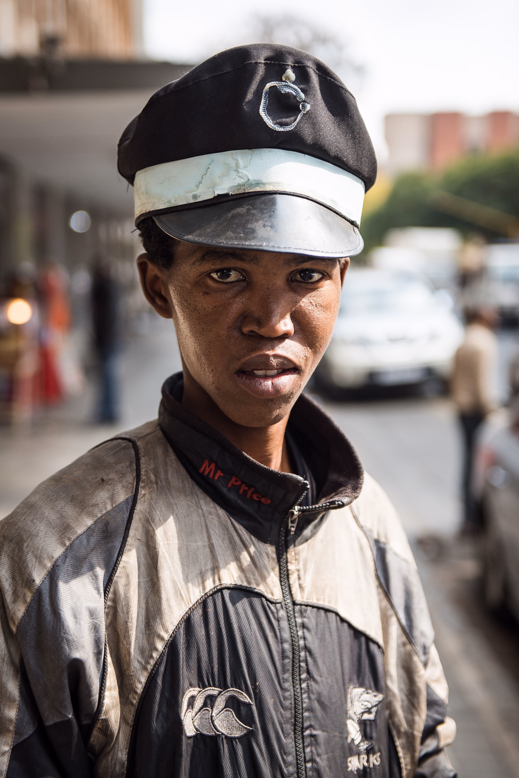 A young man in a peaked cap with a metal badge and a silver jacket looks directly at the camera on a street.
