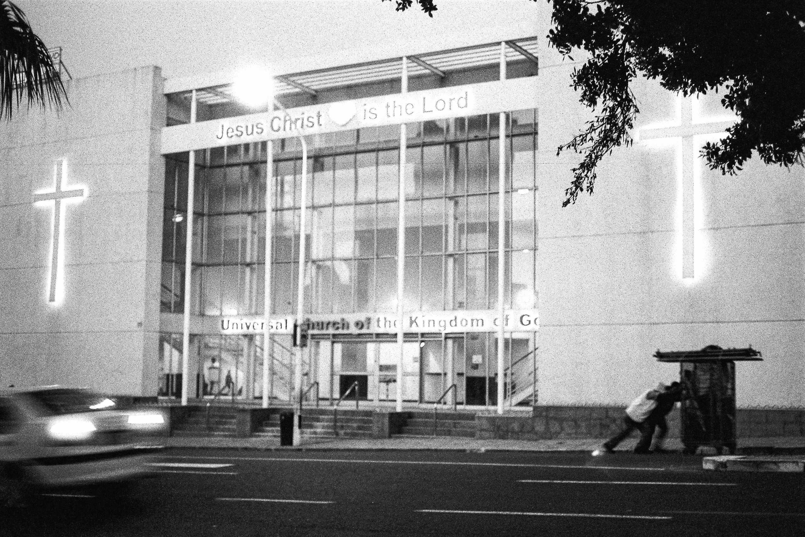 A church facade with two illuminated crosses and a lit sign, a blurred car passing and a man pushing a trolley on the right, Cape Town.