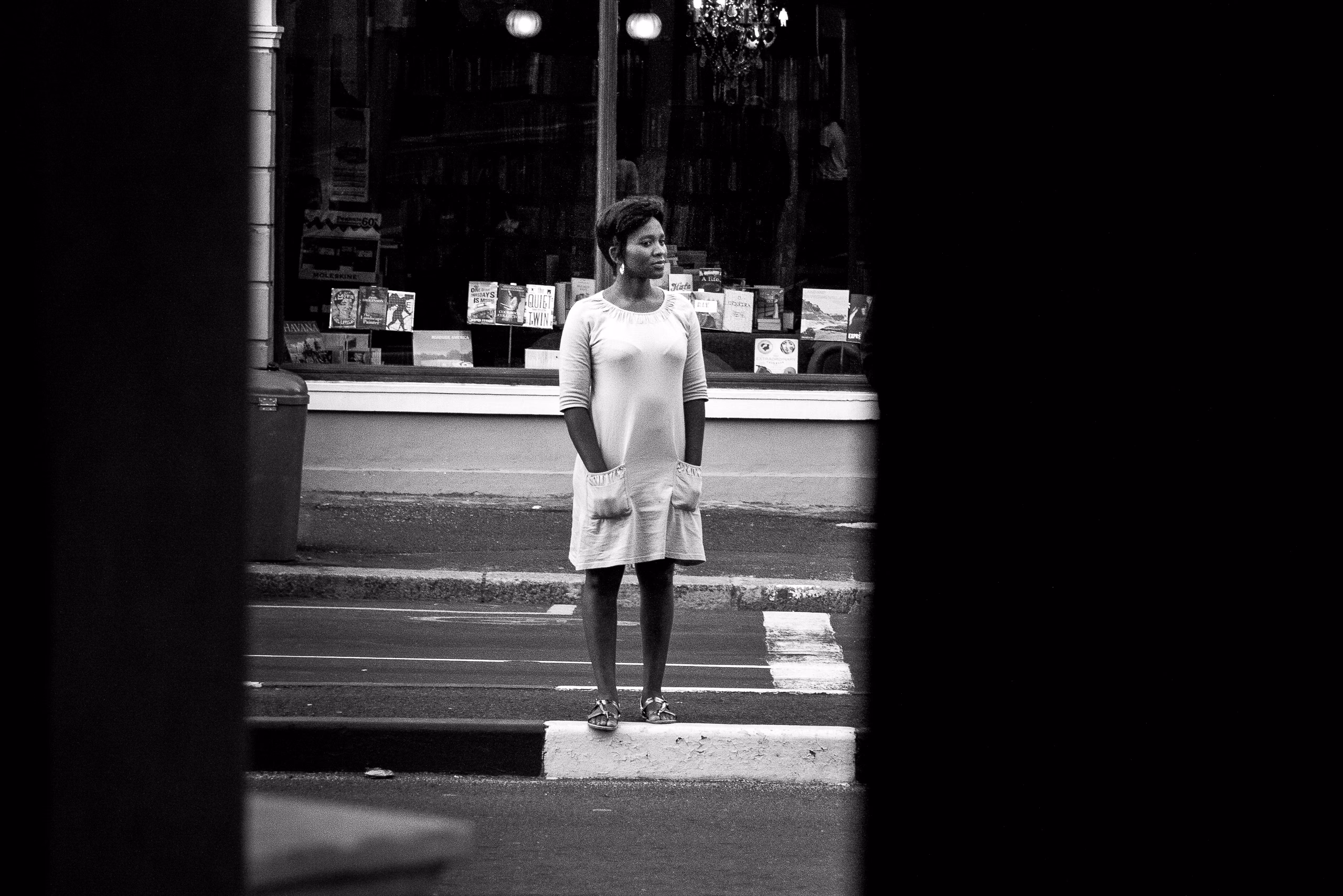 A woman in a pale dress standing on a narrow traffic island in front of a bookshop window, framed by dark vertical edges.