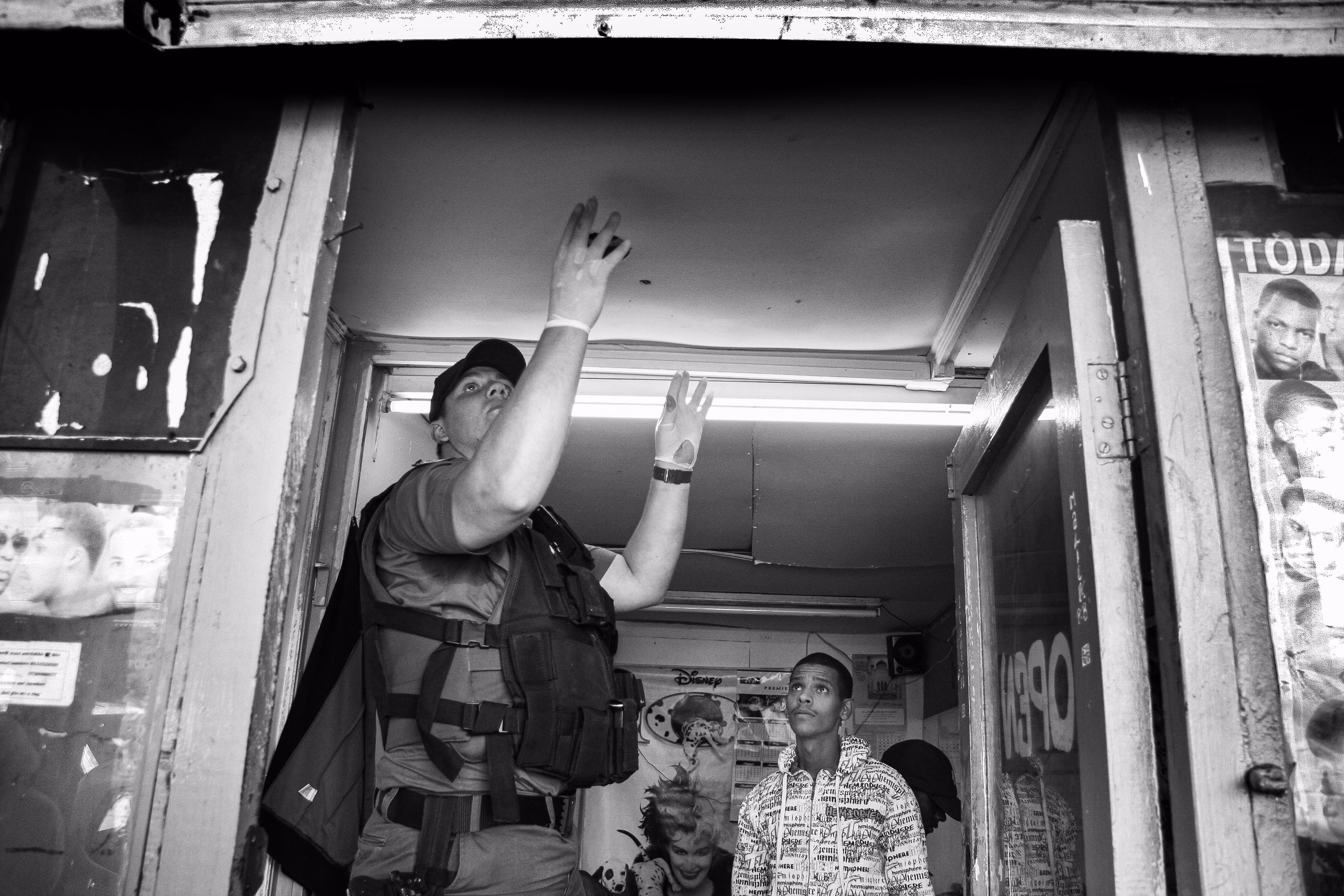 Photograph taken from below of a police officer in a tactical vest with both hands raised in a shop doorway, two men standing inside behind him.