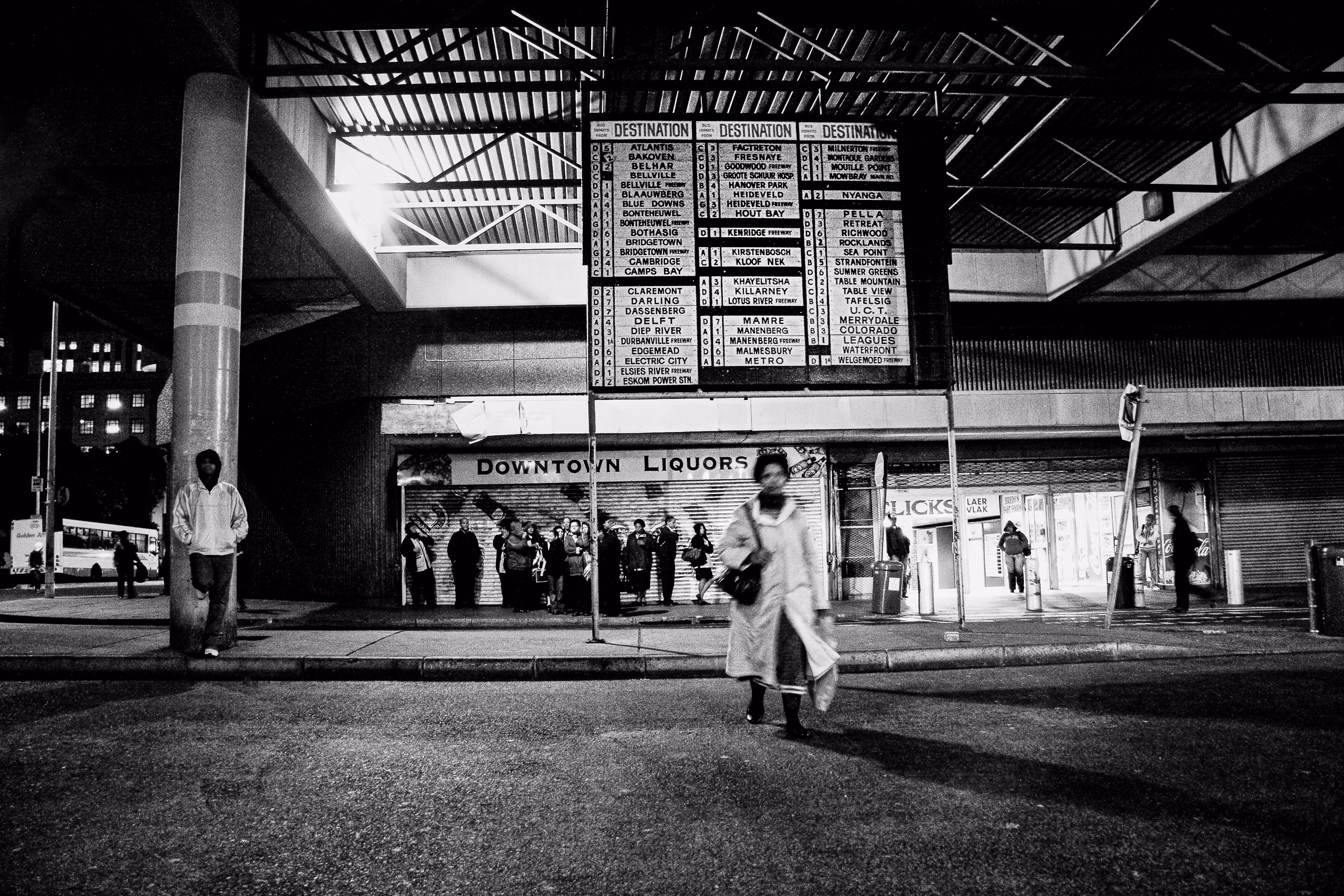Central Cape Town bus terminus and a large bus destination board listing dozens of place names, with people waiting beneath it outside shuttered shops.