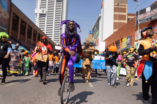 A performer in a purple and orange jester costume rides a unicycle down the middle of a city street, with a costumed parade and banners following behind.