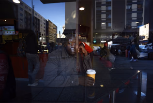 A man sits with a takeaway coffee cup behind a window, the reflected street and buildings overlaying his face and jacket.