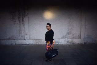 A woman in a black top and patterned skirt walks along a pavement carrying a sports bag, beneath a round pool of light on a dark wall.
